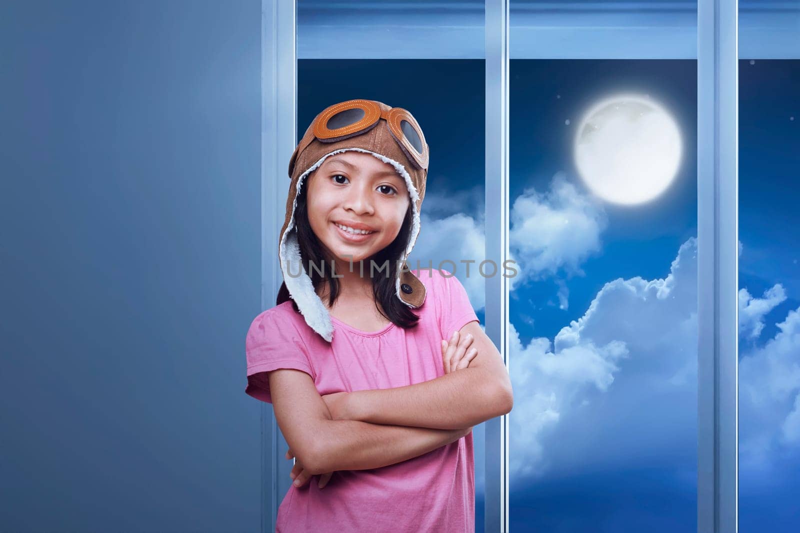 Smiling asian girl wearing goggles and aviator cap standing in her room at night