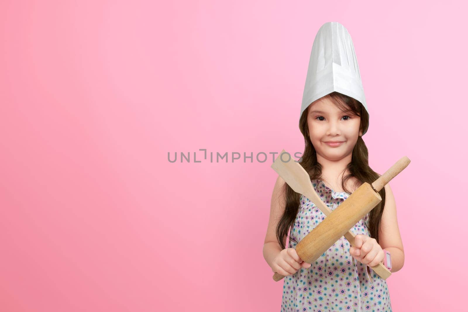 Smiling asian little girl in chef hat holding wooden cooking utensils over pink background
