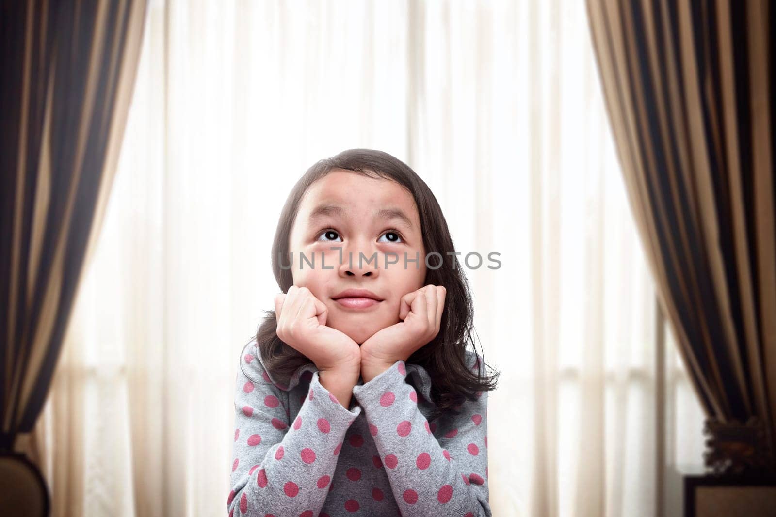 Beautiful little asian girl thinking and daydreaming at living room in front of the window