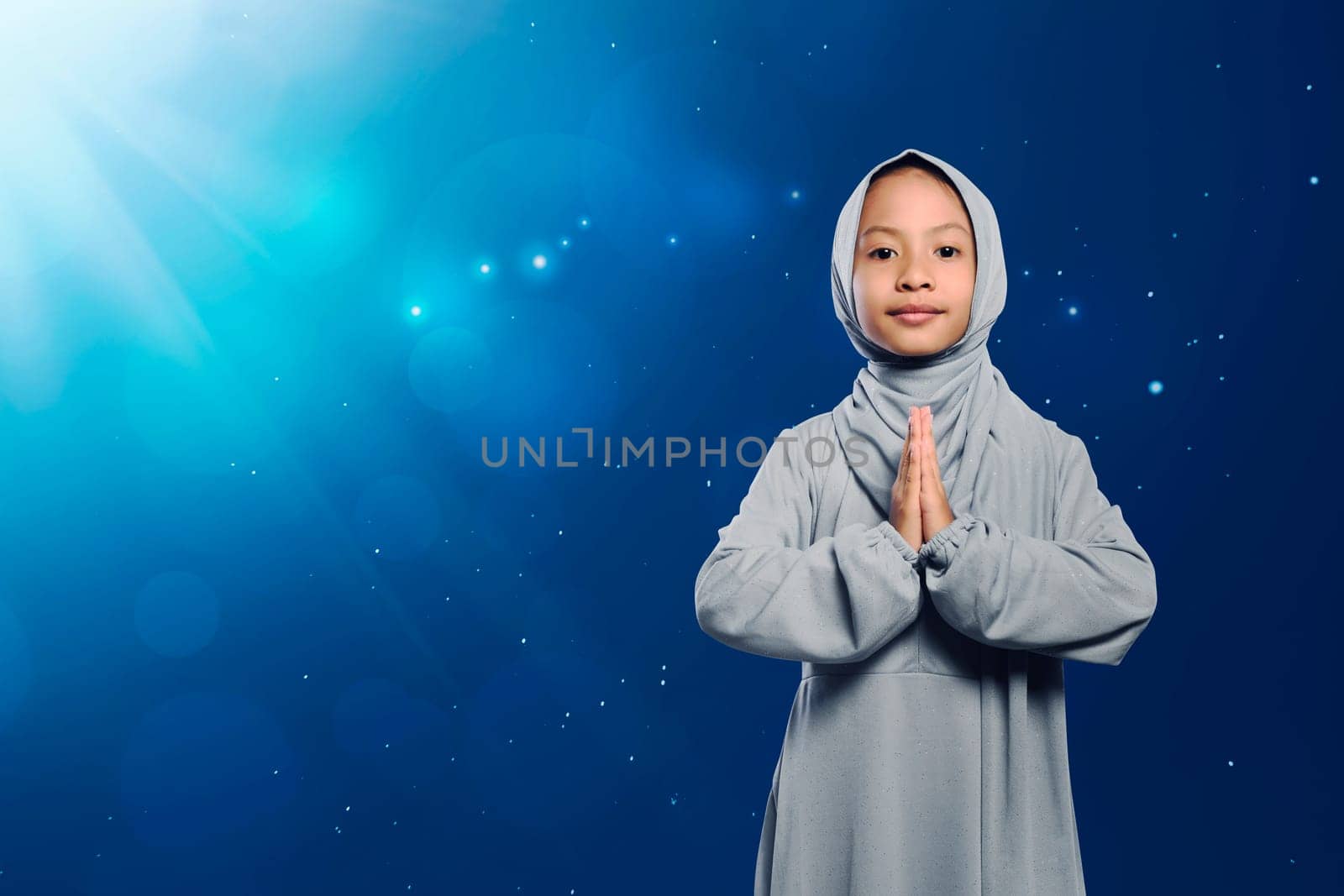 Little asian muslim girl in traditional dress praying against light background