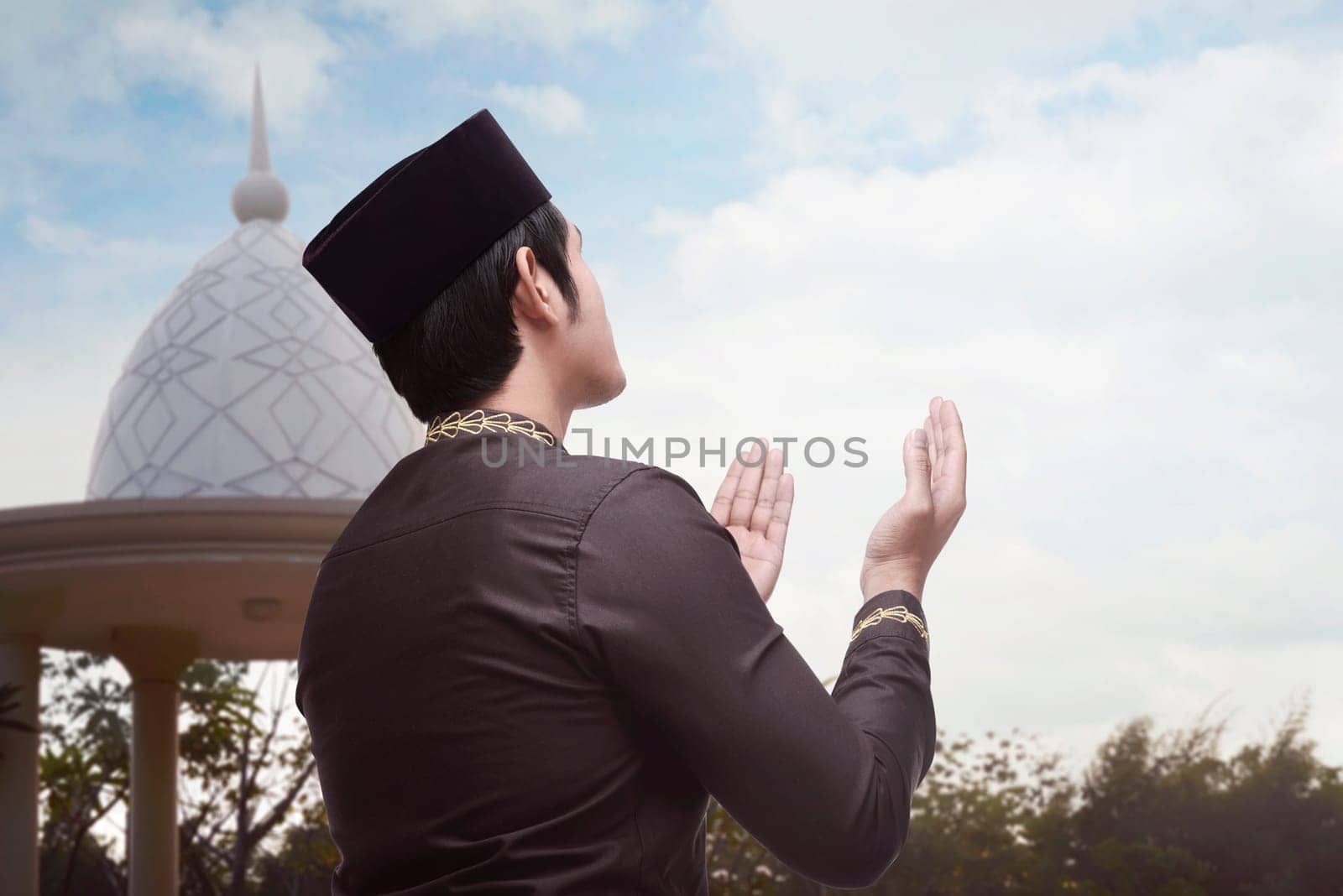 Back view of asian muslim man raising both his hand to pray with blue sky background