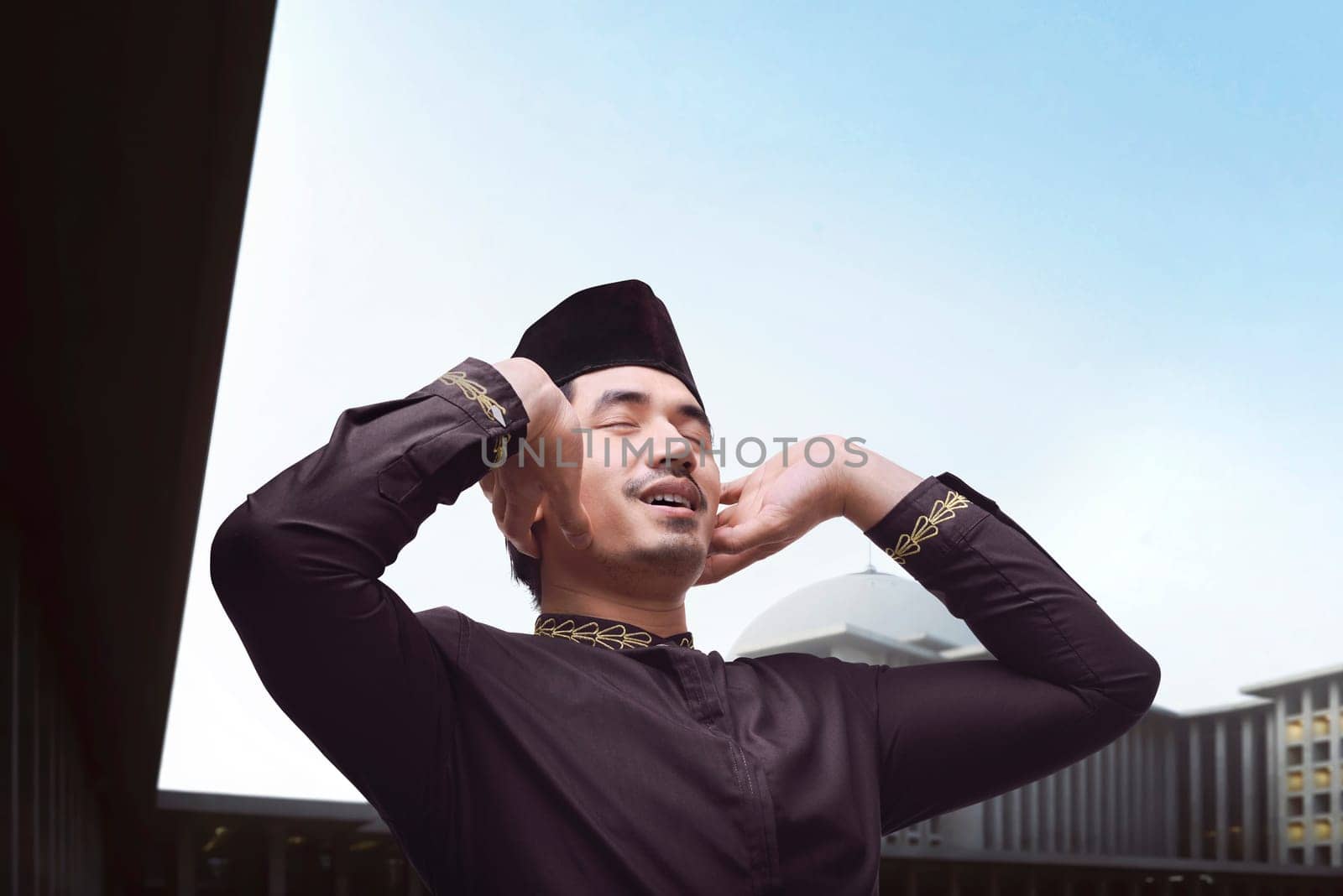 Handsome asian muslim man in traditional dress praying with mosque background