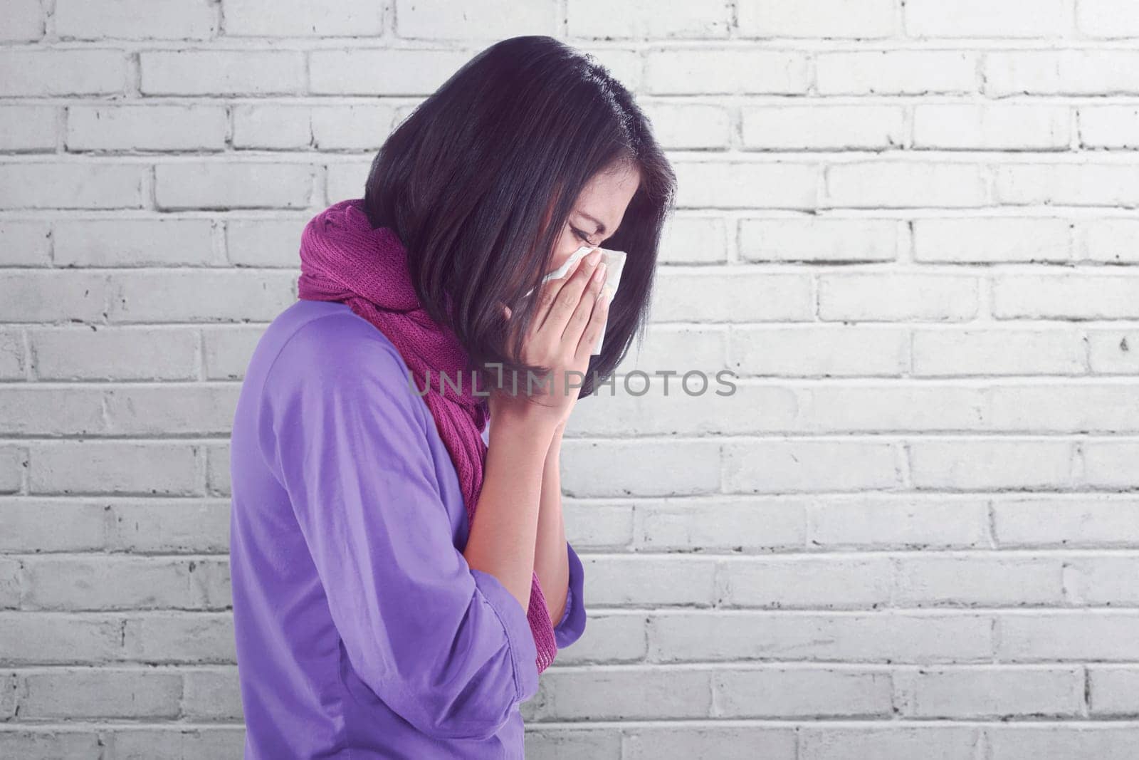Sick asian woman with flu and fever holding tissue in nose against wall background