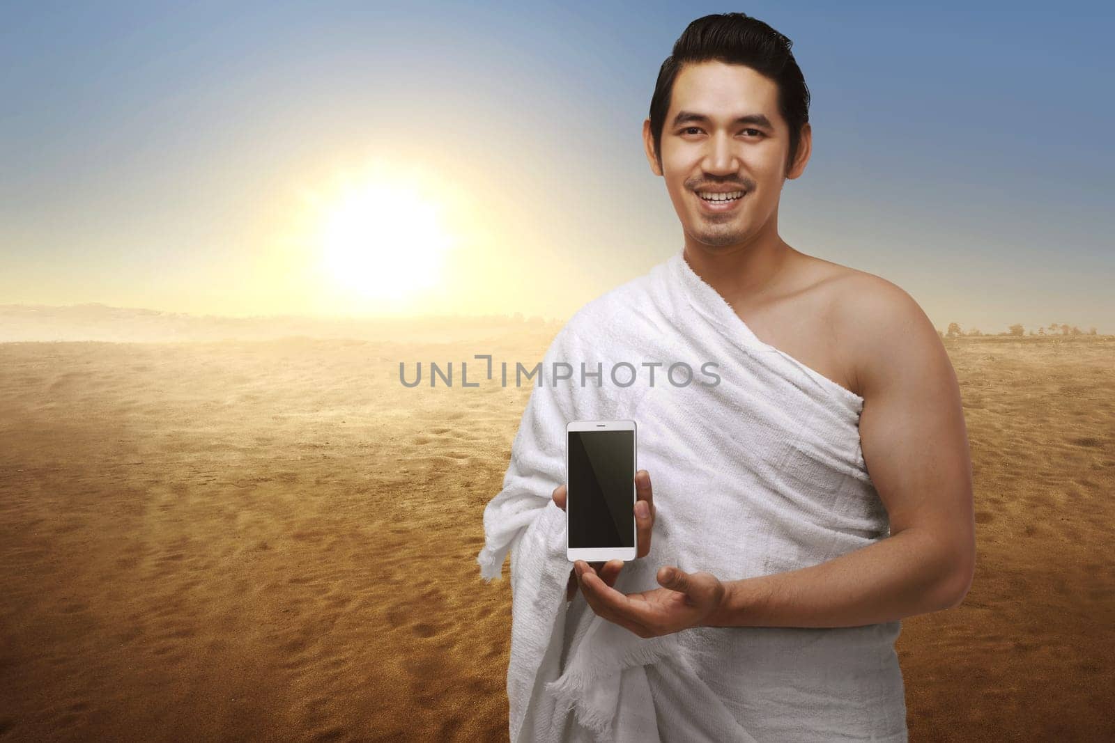 Handsome asian muslim man with ihram clothes showing phone against sunset background