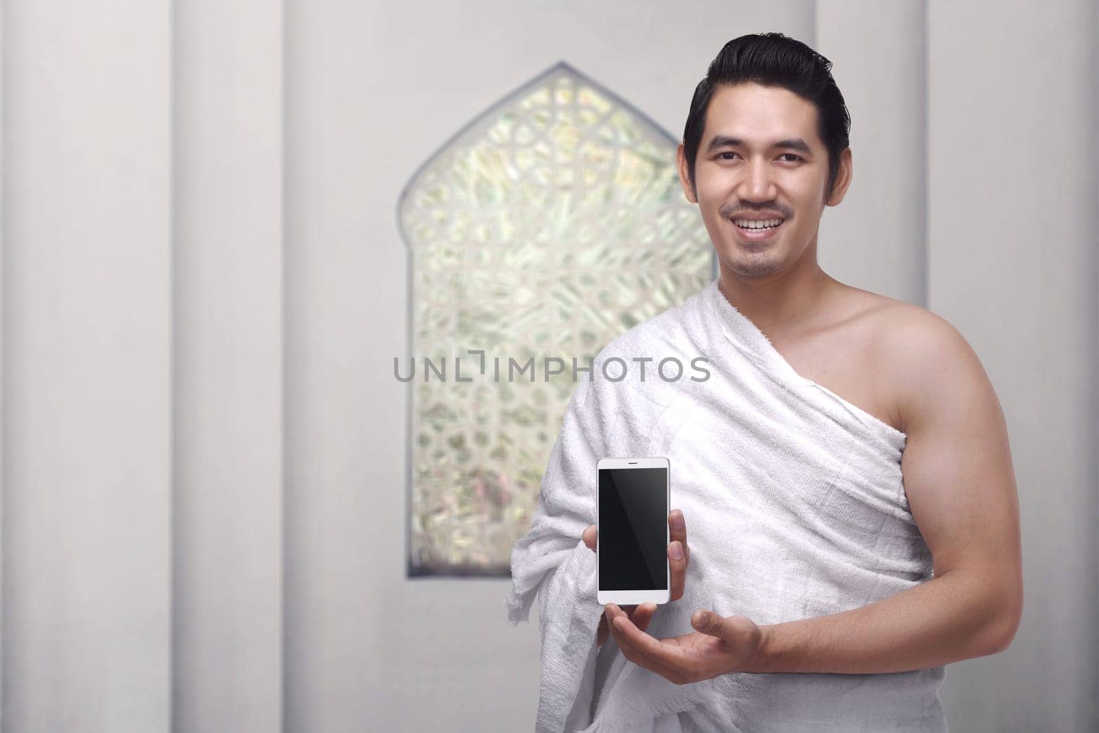 Handsome asian muslim man wearing ihram clothes holding mobile phone against white wall background