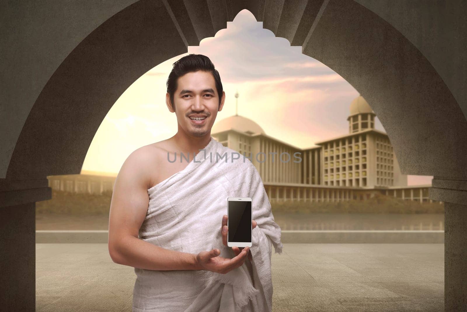 Cheerful asian muslim man with ihram clothes show mobile phone with mosque background