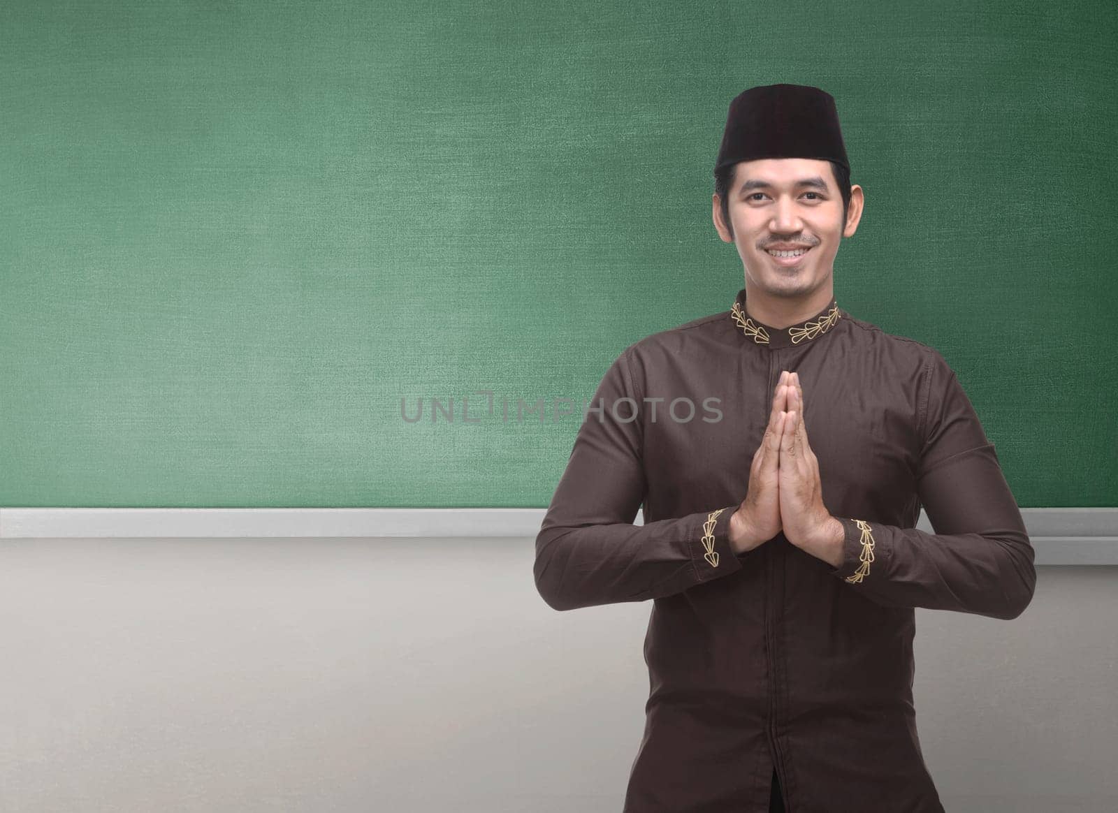 Cheerful asian muslim man wearing traditional dress with hand gesture against chalkboard background for copyspace