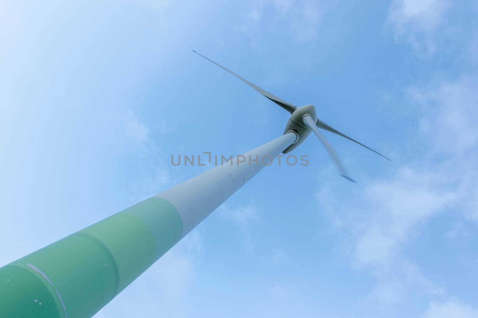 Individual green-and-white striped wind turbine in extreme inclination upwards in front of slightly cloudy skies.