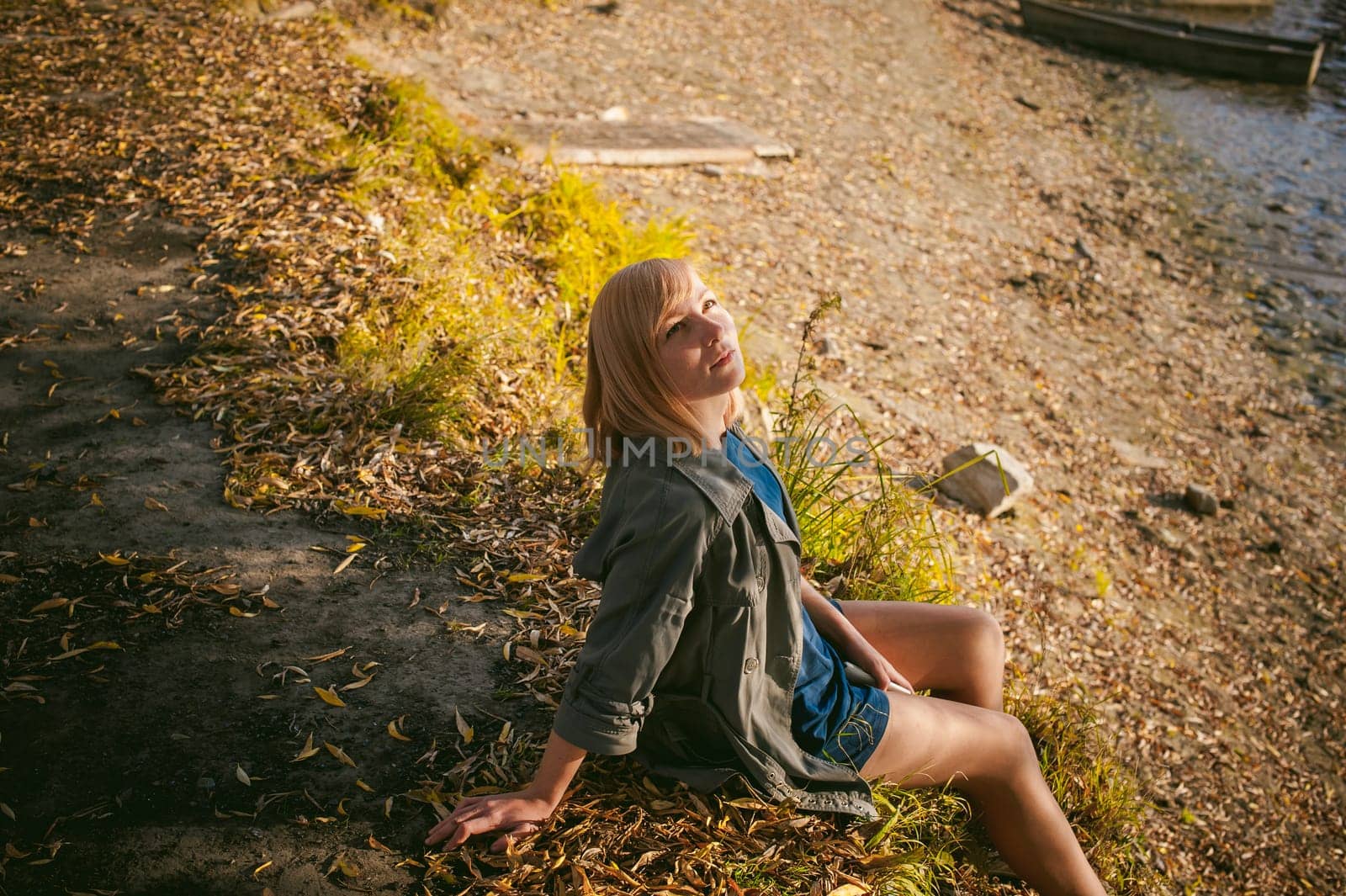 pretty blonde girl in sneakers, nylon stockings and coat, sits on  banks of river, on yellow leaves, enjoying warmth of autumn sun rays