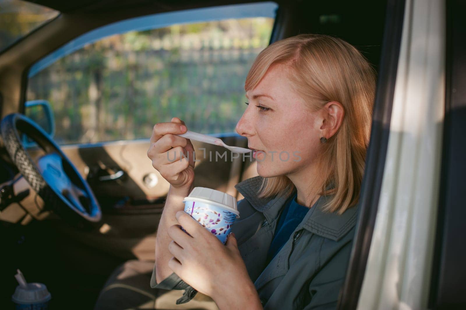 girl eating ice cream with a spoon in the car. blond girl in a raincoat, eats ice cream while in the car during a road trip