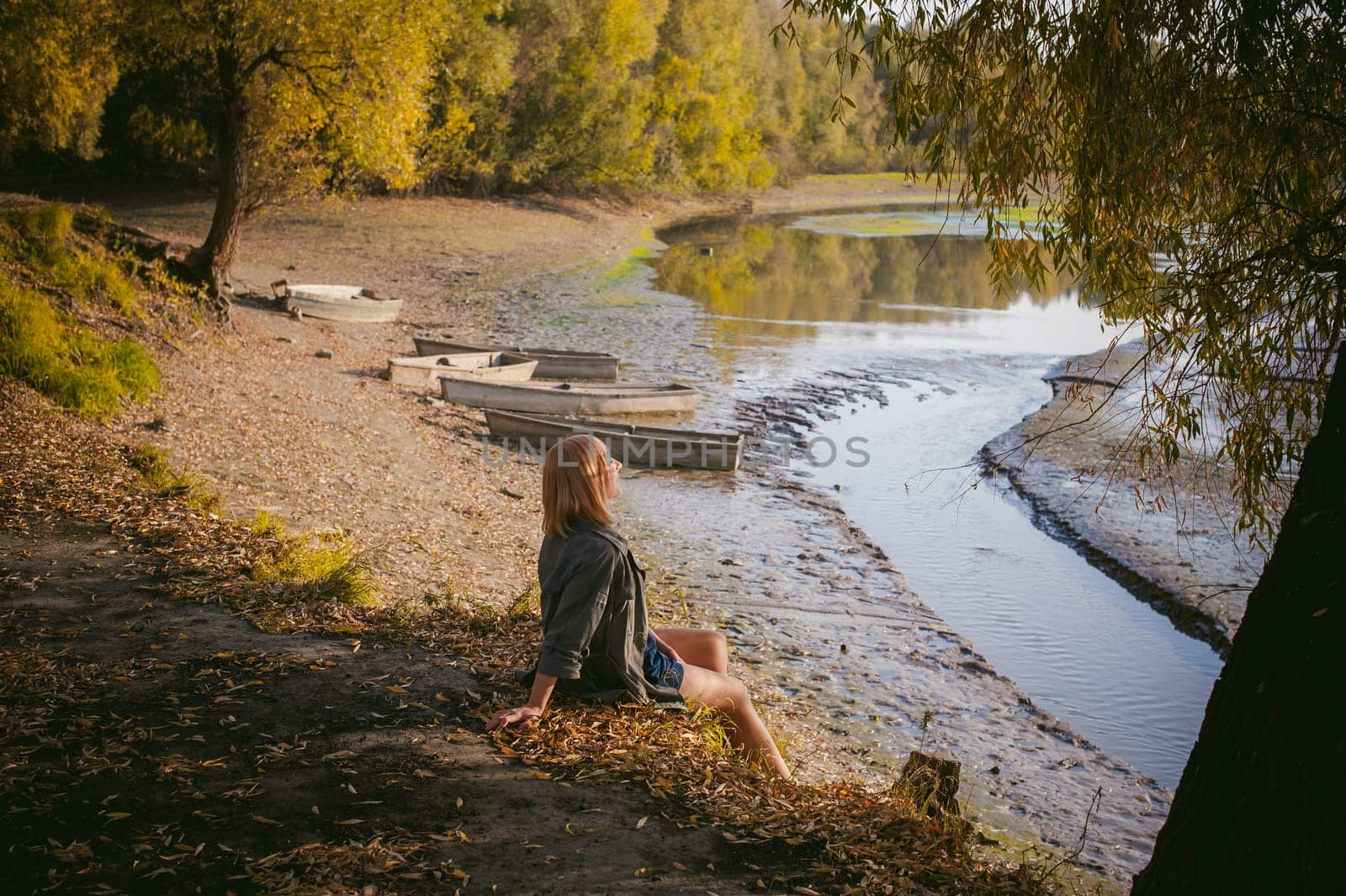pretty blonde girl in sneakers, nylon stockings and coat, sits on  banks of river, on yellow leaves, enjoying warmth of autumn sun rays