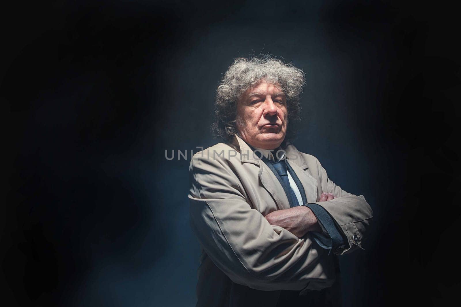 The senior man in cloak standing as detective or mafia boss. Studio shot on gray in retro stile in the light and smoke ray