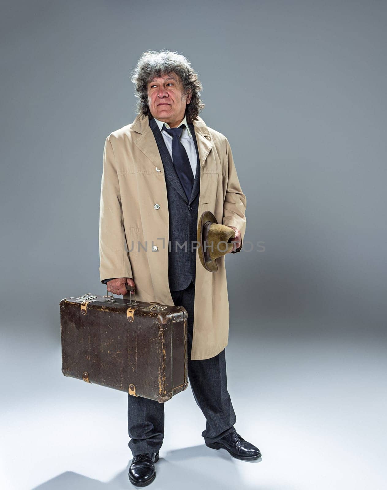 The senior man in cloak as detective or mafia boss. Studio shot on gray in retro stile. Mature man with hat and suitcase