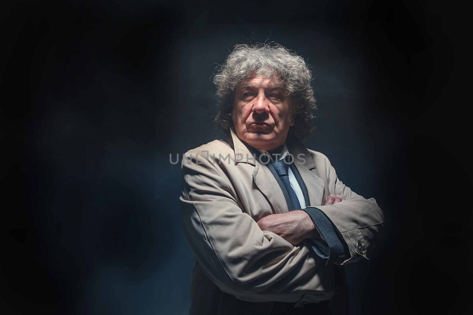 The senior man in cloak standing as detective or mafia boss. Studio shot on gray in retro stile in the light and smoke ray