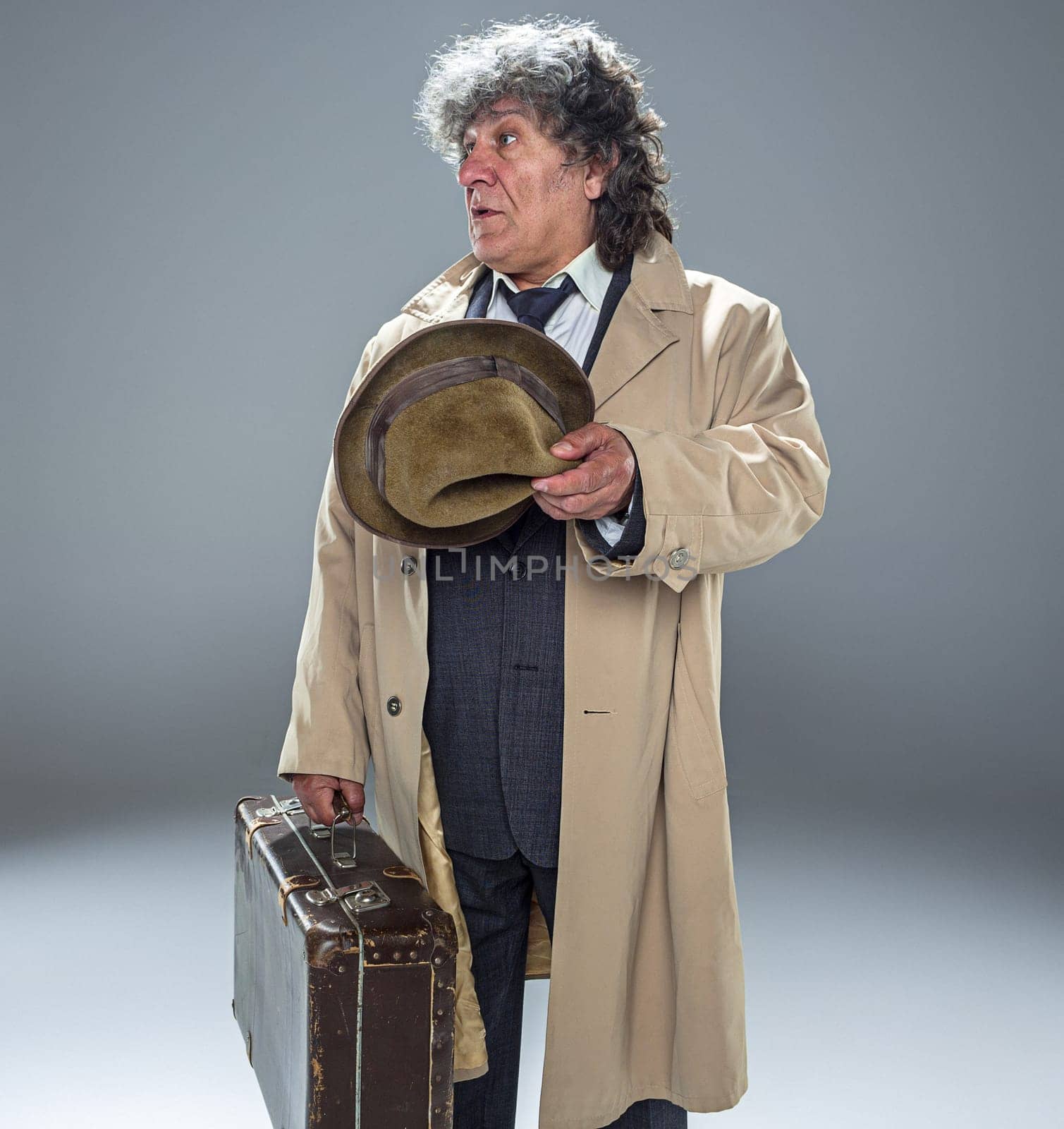 The senior man in cloak as detective or mafia boss. Studio shot on gray in retro stile. Mature man with hat and suitcase