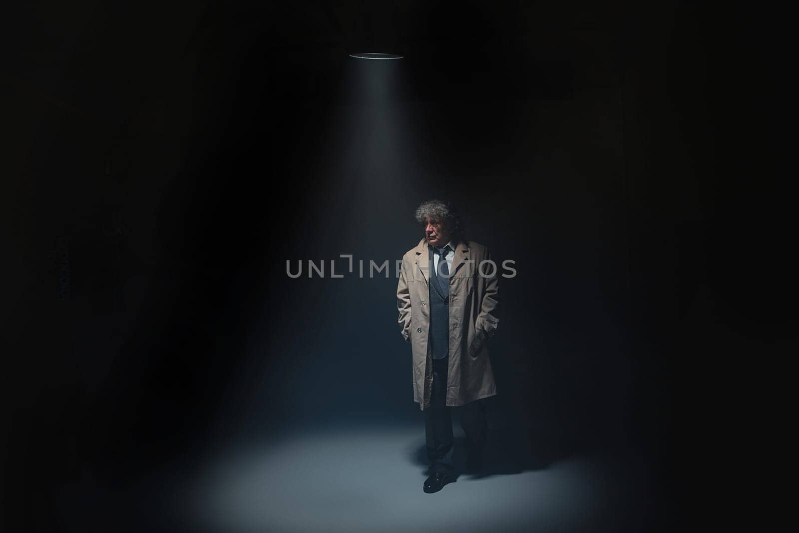The senior man in cloak standing as detective or mafia boss. Studio shot on gray in retro stile in the light and smoke ray