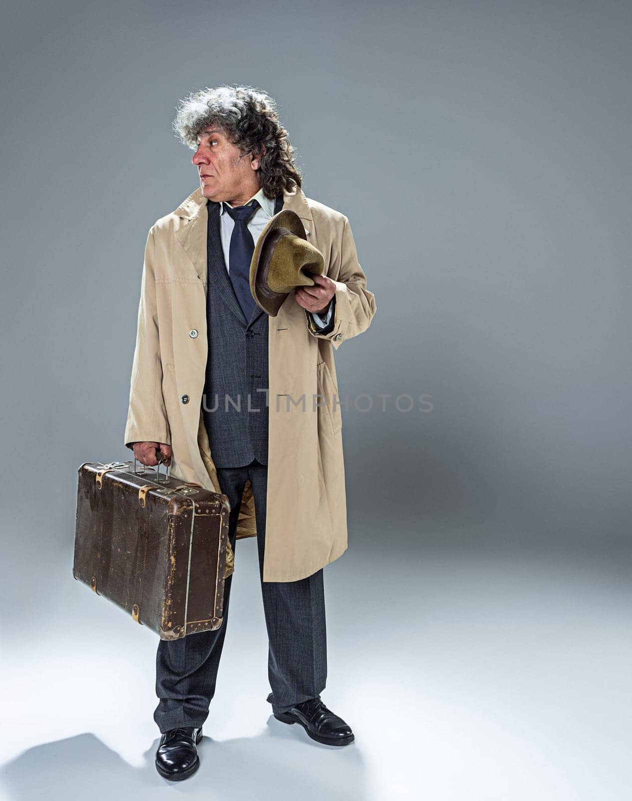 The senior man in cloak as detective or mafia boss. Studio shot on gray in retro stile. Mature man with hat and suitcase