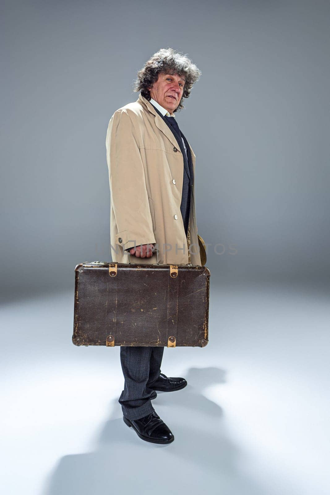 The senior man in cloak as detective or mafia boss. Studio shot on gray in retro stile. Mature man with hat and suitcase