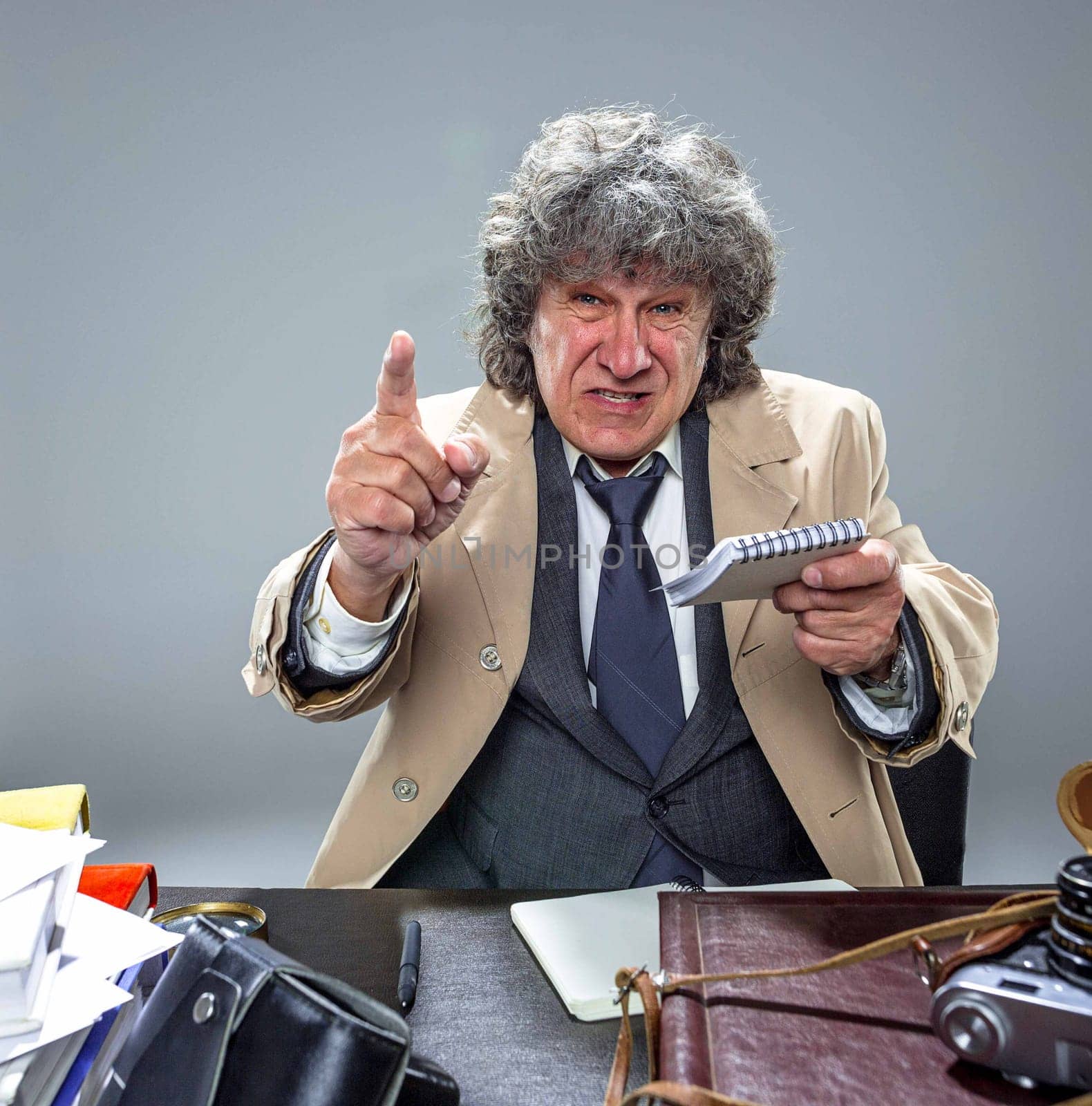 The senior man in cloak as detective or mafia boss pointing at camera at table. Studio shot on gray in retro stile