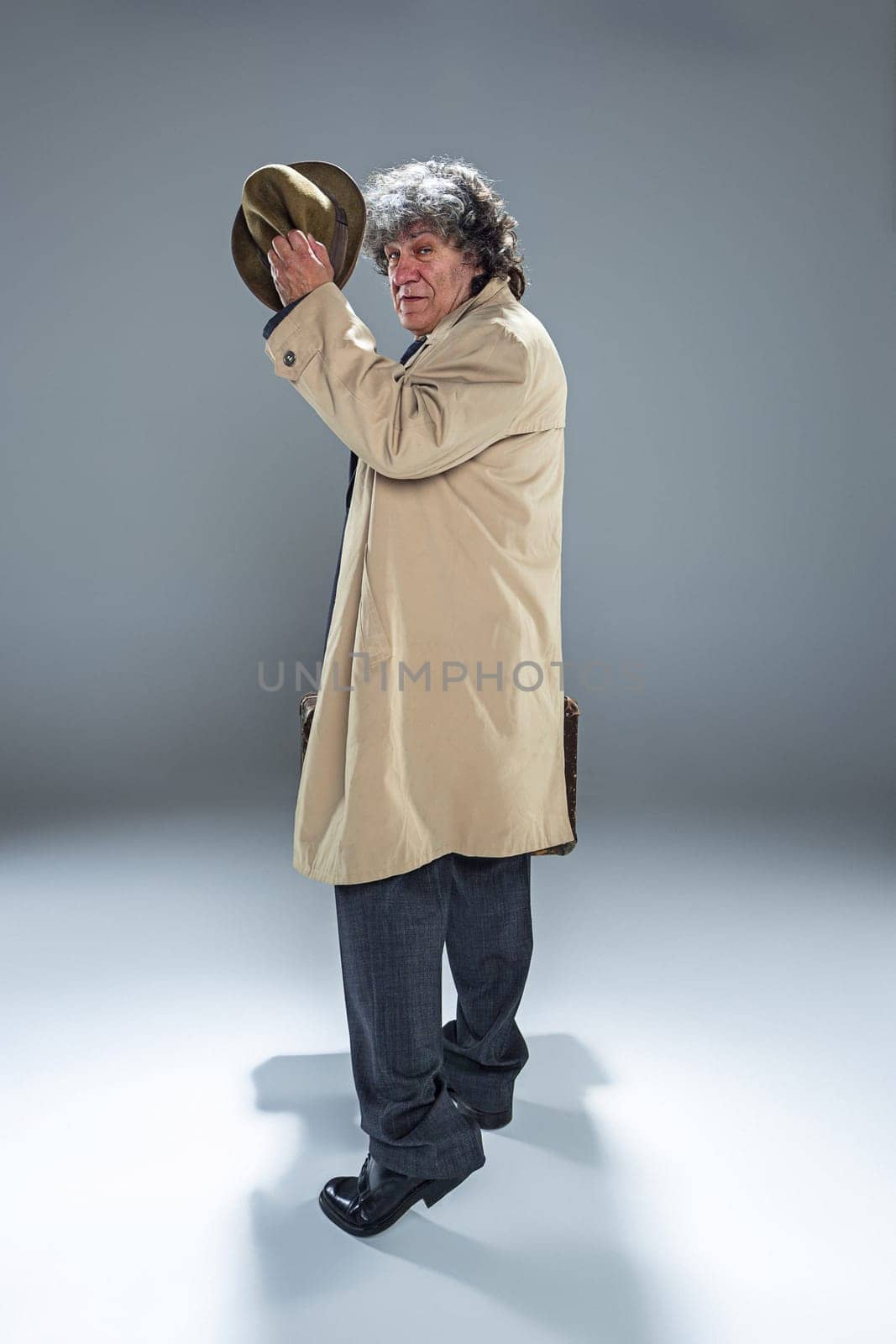 The senior man in cloak as detective or mafia boss. Studio shot on gray in retro stile. Mature man with hat and suitcase