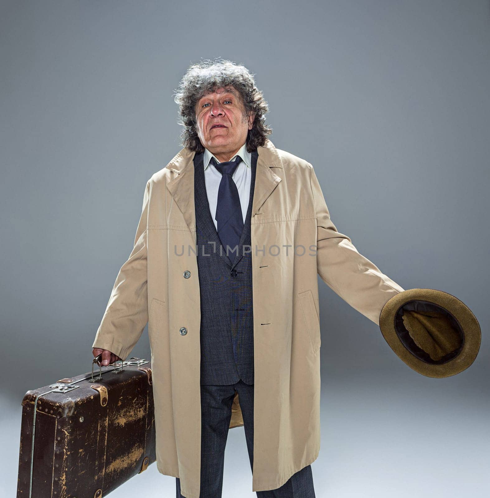 The senior man in cloak as detective or mafia boss. Studio shot on gray in retro stile. Mature man with hat and suitcase