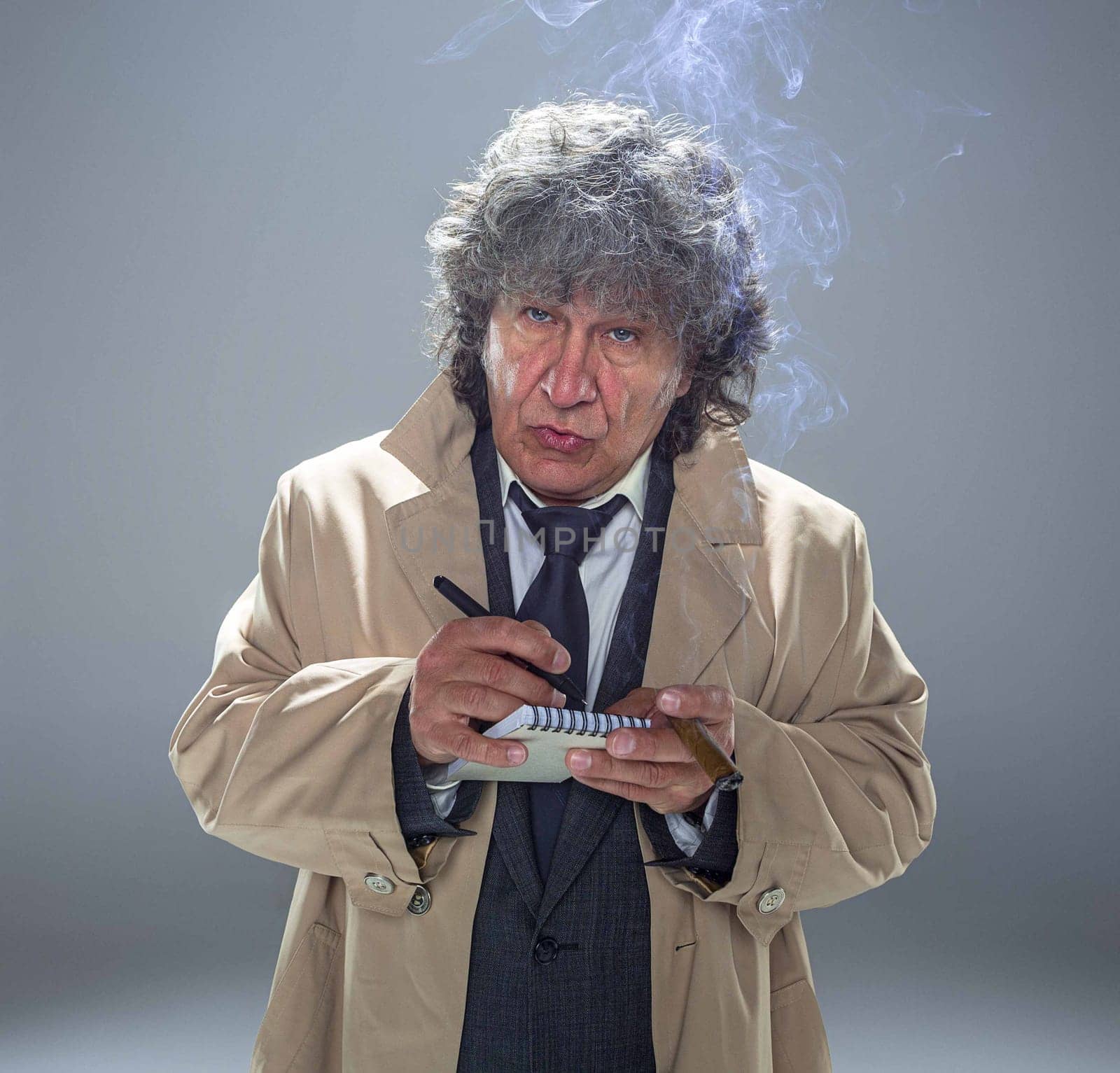 The senior man in cloak as detective or mafia boss writing in a notebook with pen. Studio shot on gray in retro stile