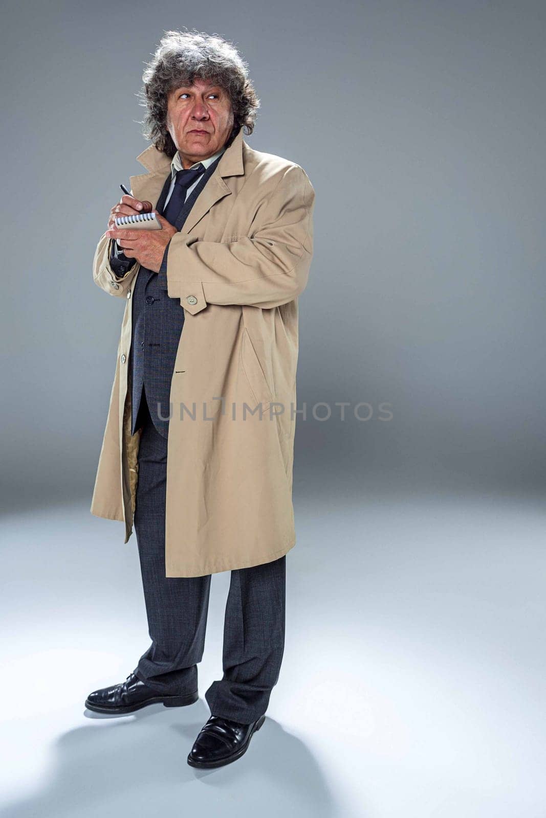 The senior man in cloak as detective or mafia boss writing in a notebook with pen. Studio shot on gray in retro stile