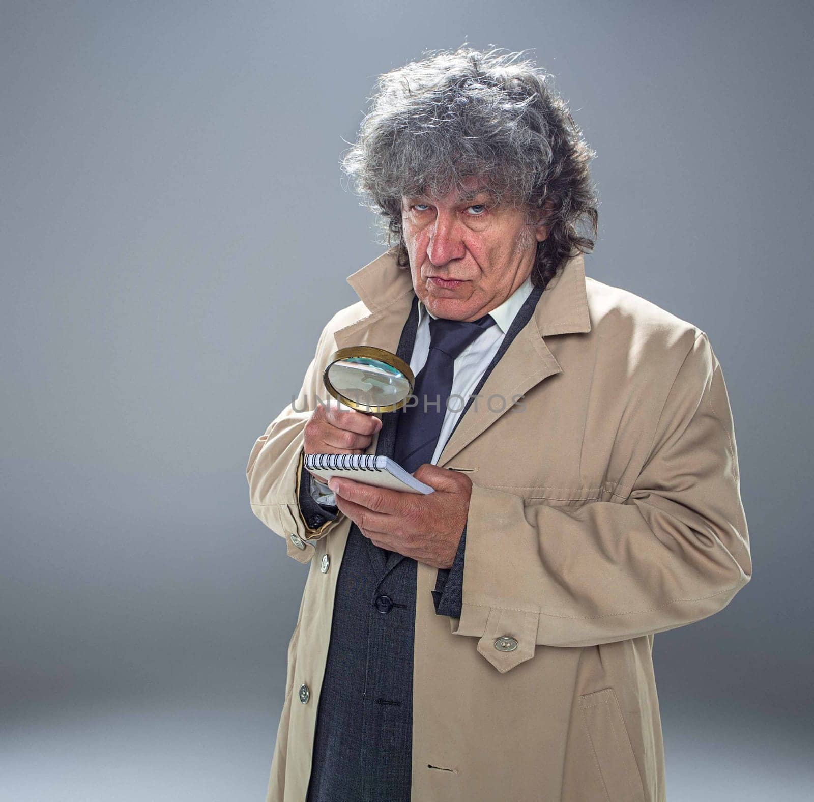 The senior man in cloak as detective or mafia boss with With a magnifying glass. Studio shot on gray in retro stile. Concept of Search