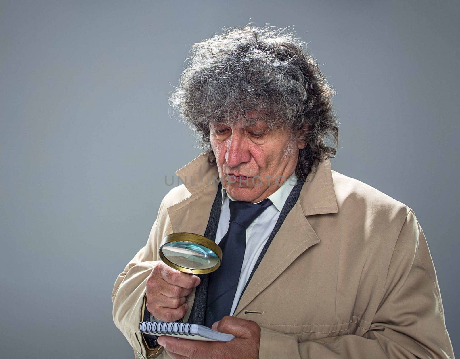 The senior man in cloak as detective or mafia boss with With a magnifying glass. Studio shot on gray in retro stile. Concept of Search