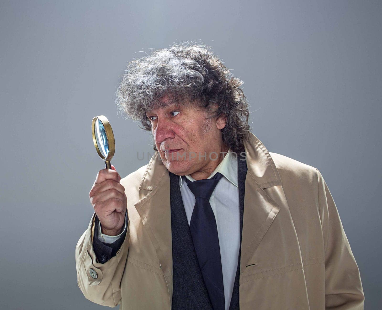 The senior man in cloak as detective or mafia boss with With a magnifying glass. Studio shot on gray in retro stile. Concept of Search