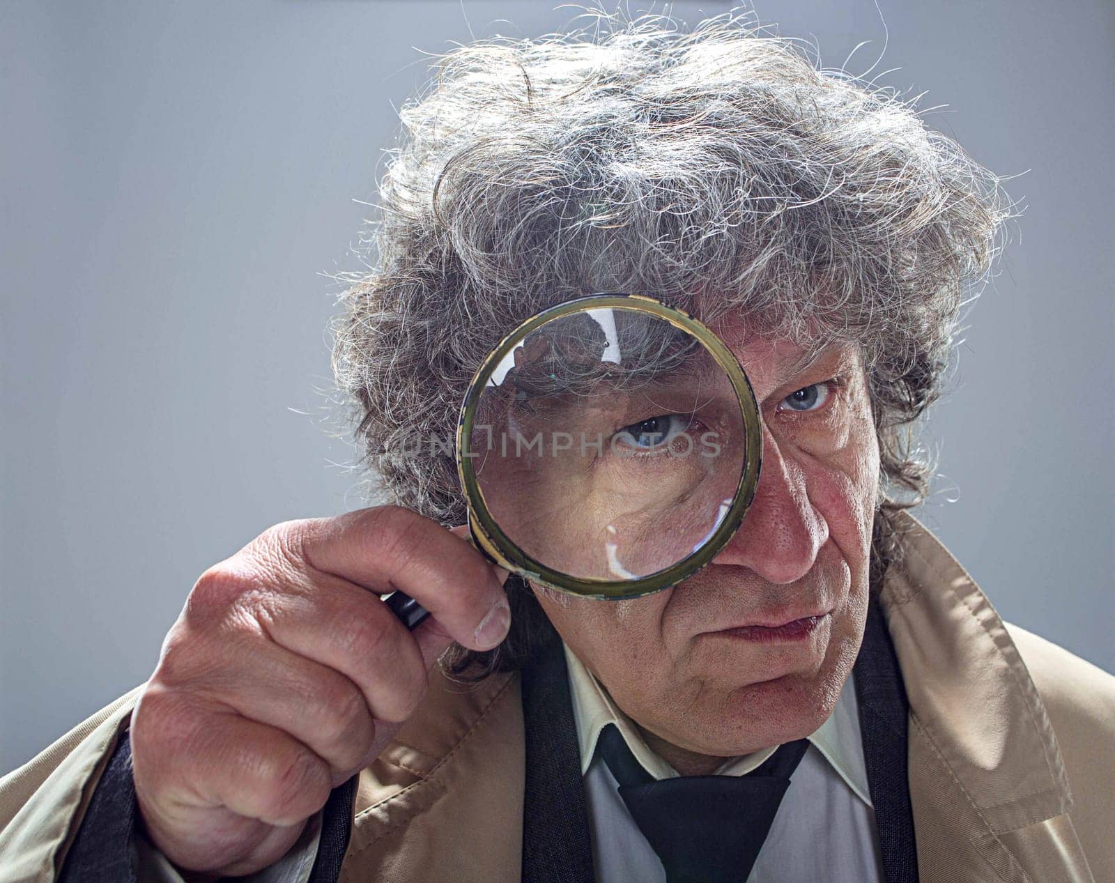 The senior man in cloak as detective or mafia boss with With a magnifying glass. Studio shot on gray in retro stile. Concept of Search