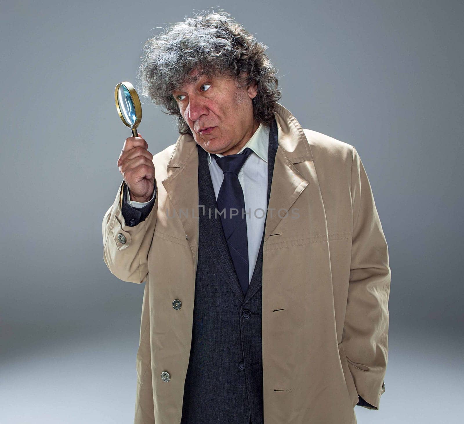 The senior man in cloak as detective or mafia boss with With a magnifying glass. Studio shot on gray in retro stile. Concept of Search