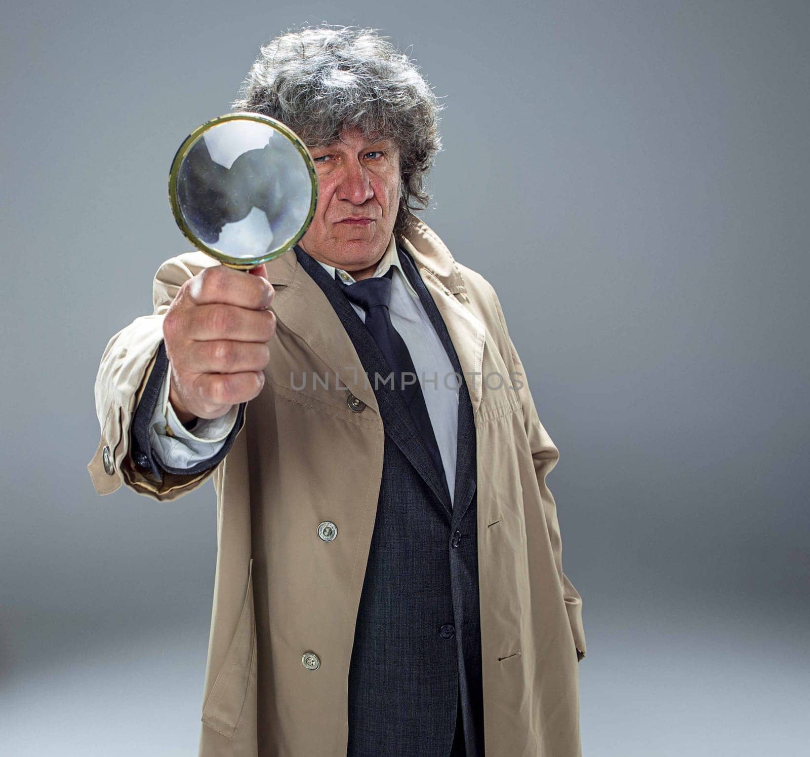 The senior man in cloak as detective or mafia boss with With a magnifying glass. Studio shot on gray in retro stile. Concept of Search