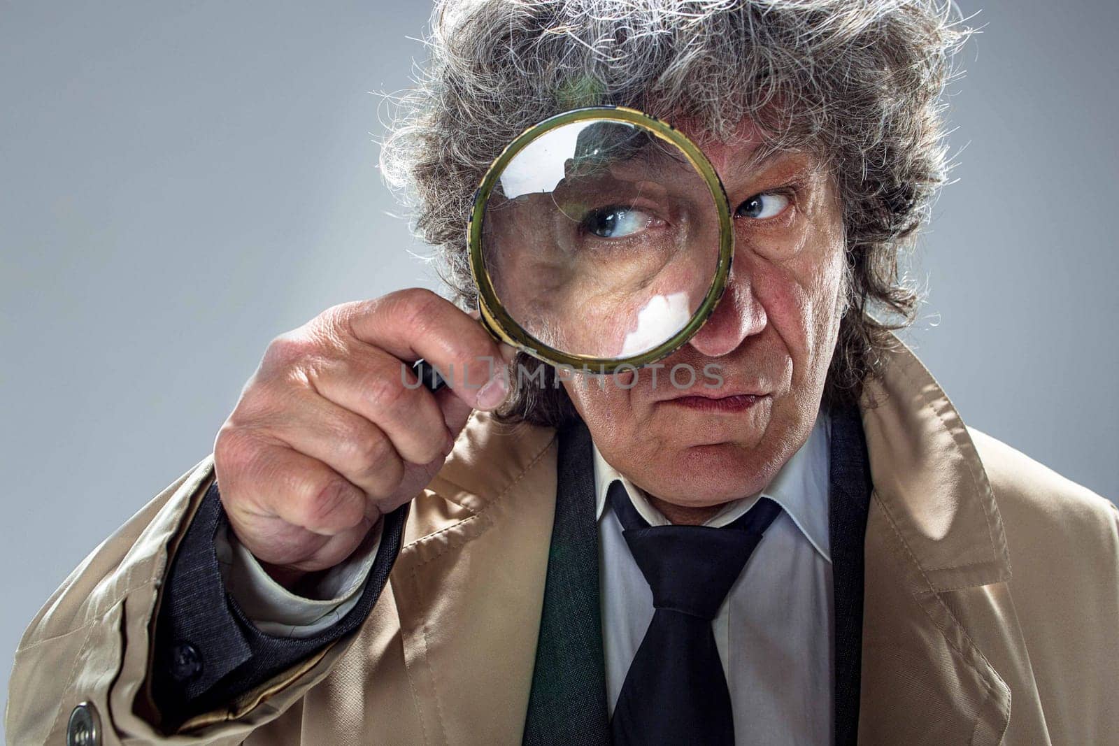 The senior man in cloak as detective or mafia boss with With a magnifying glass. Studio shot on gray in retro stile. Concept of Search