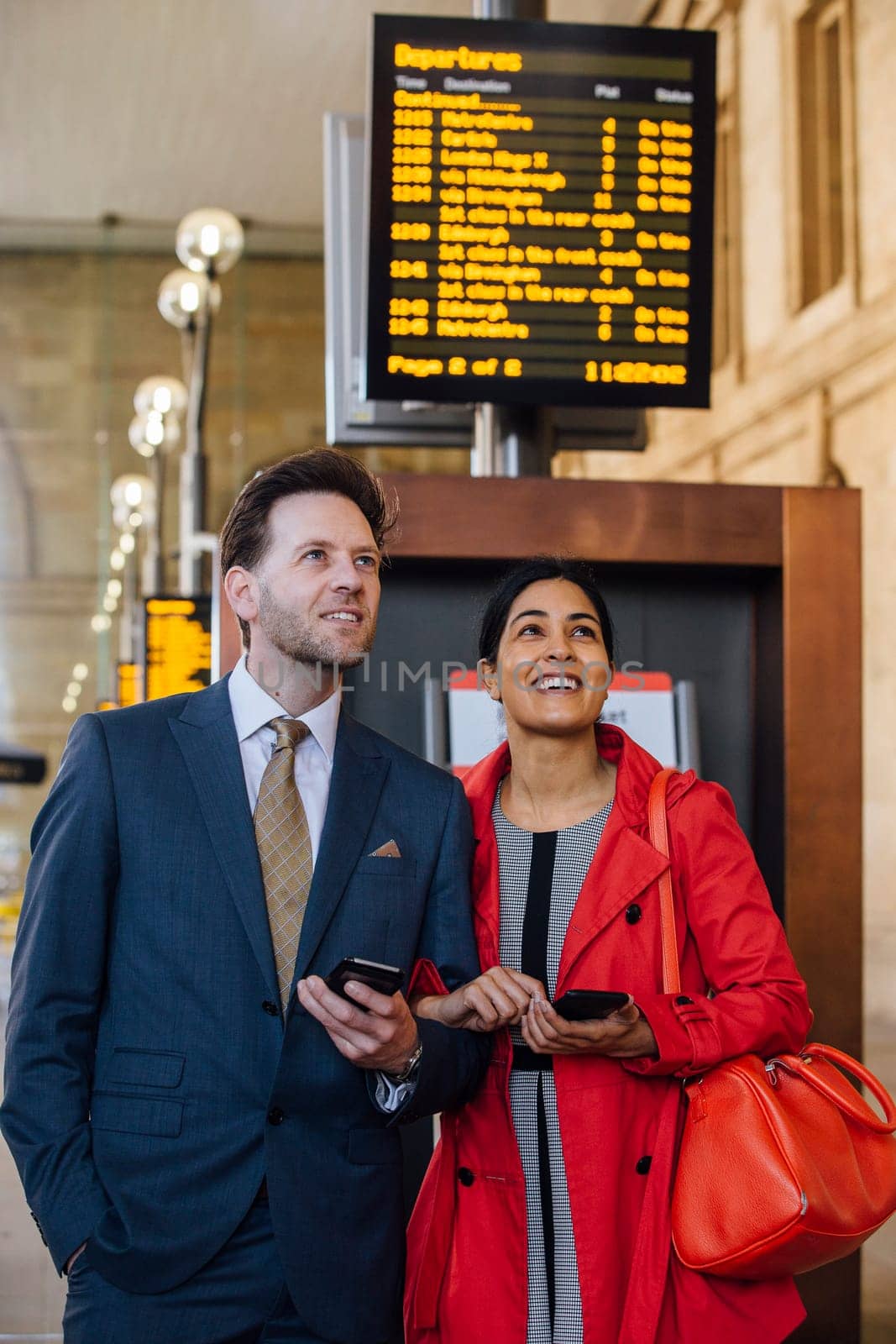 Couple are standing together in the train station with their smart phones in hand, looking up at a board of train times.