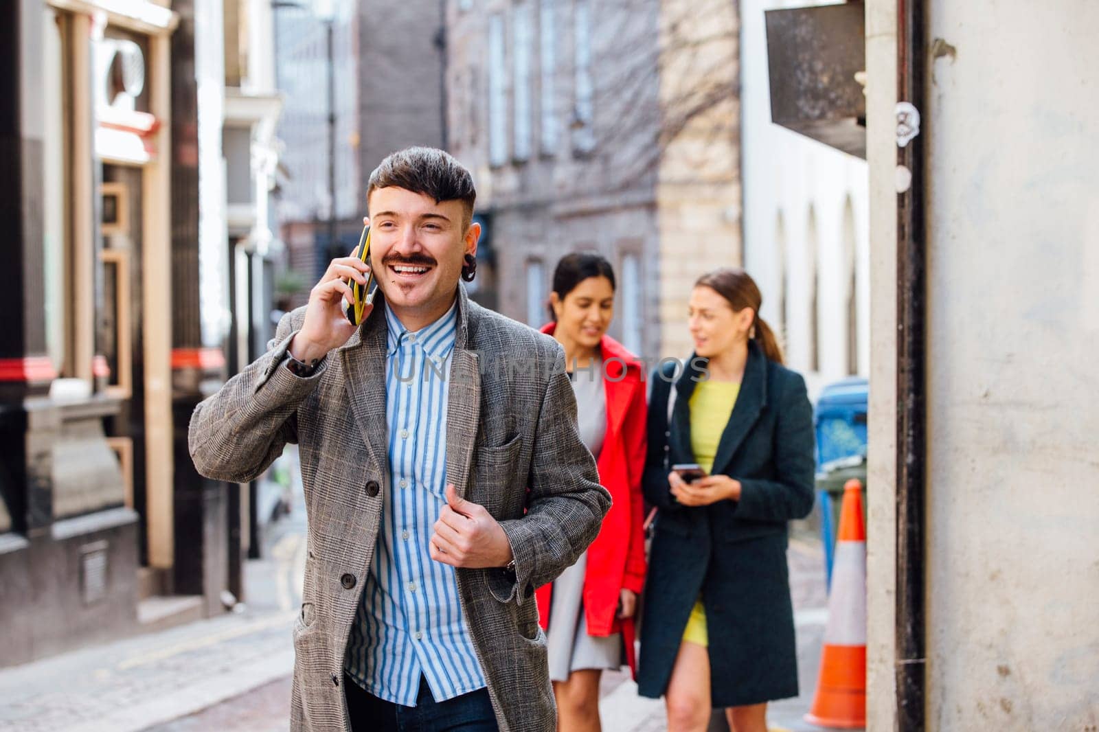 Businessman is walking through the city to go to work. He is well dressed and is talking on the phone, while two women can be seen in the background walking together while lookin at a smart phone.
