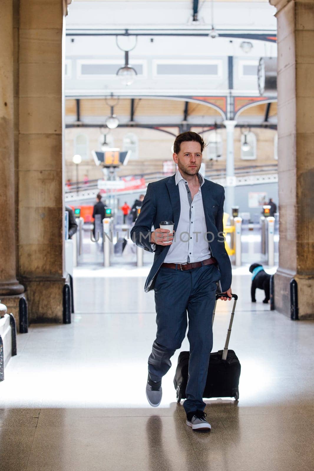 Businessman is in a hurry as he leaves the train station to commute the work. He is carrying a coffee and pulling a suitcase.