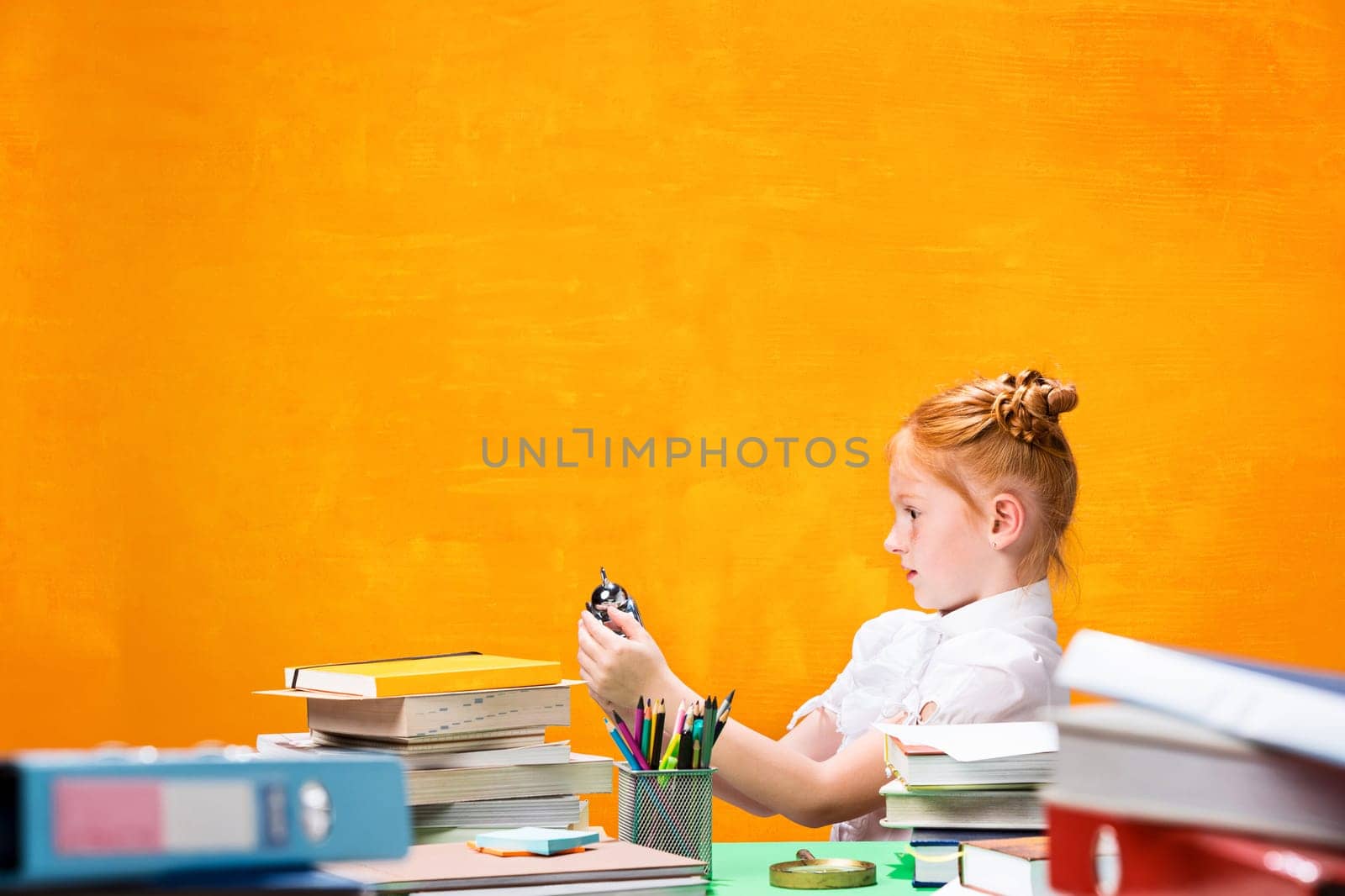 The teen girl with lot of books sitting with Magnifier at table on orange studio background. The education and back to school concept