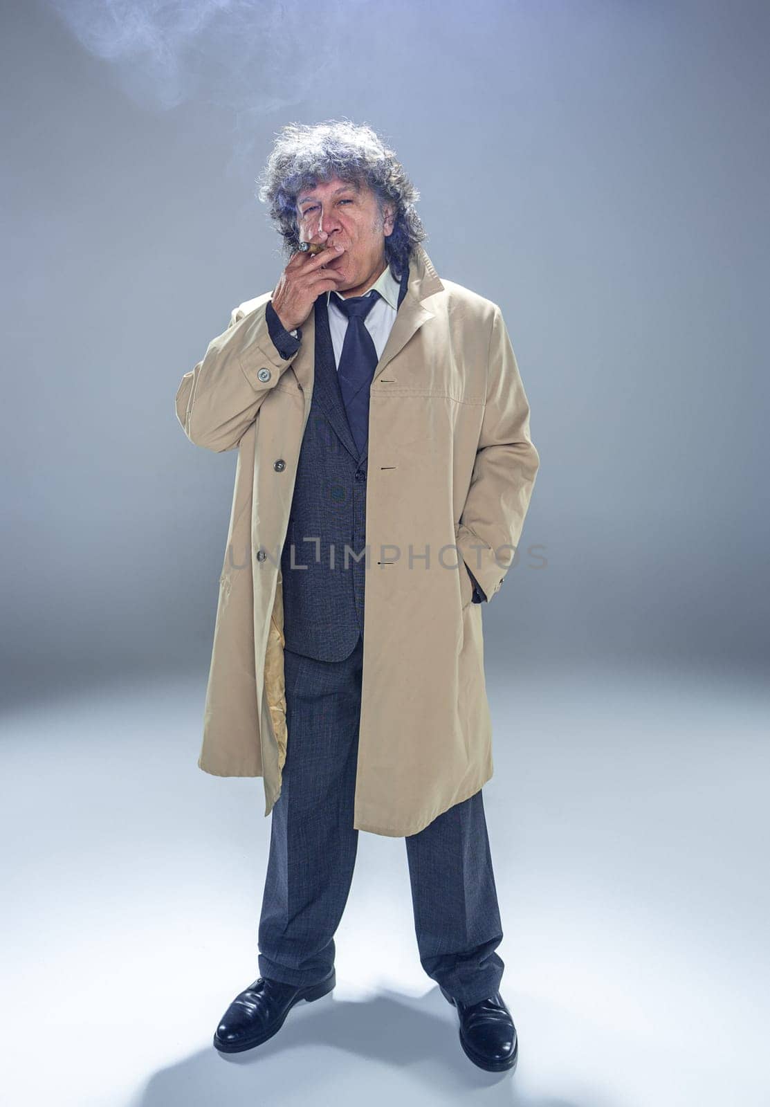 The senior man in cloak with cigar as detective or mafia boss. Studio shot on gray in retro stile