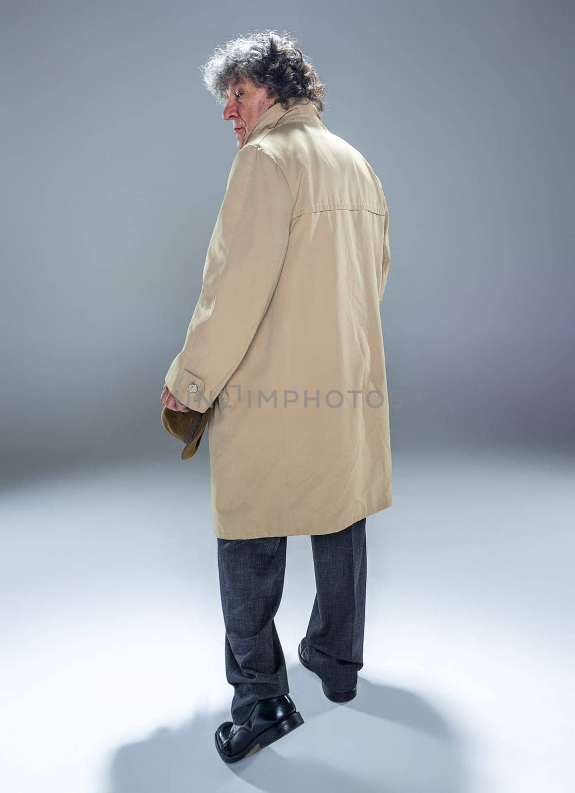 The back view of senior man in cloak as detective or mafia boss. Studio shot on gray in retro stile. Mature man with hat and suitcase
