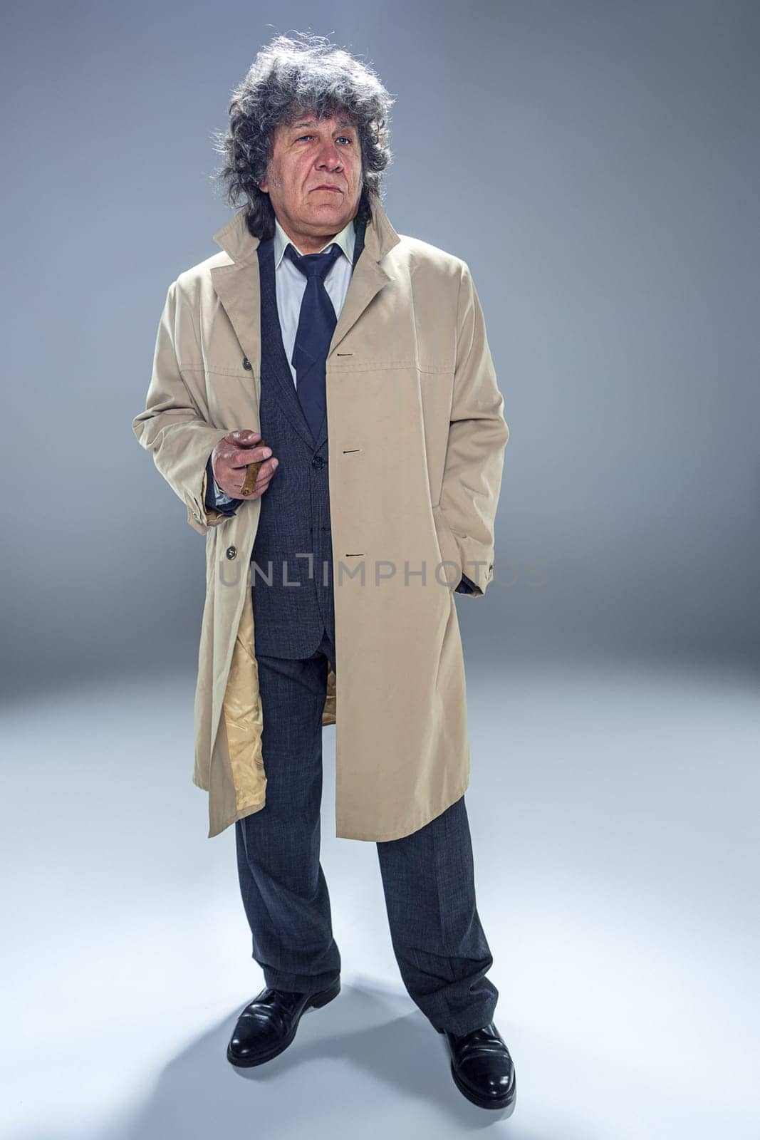The senior man in cloak with cigar as detective or mafia boss. Studio shot on gray in retro stile