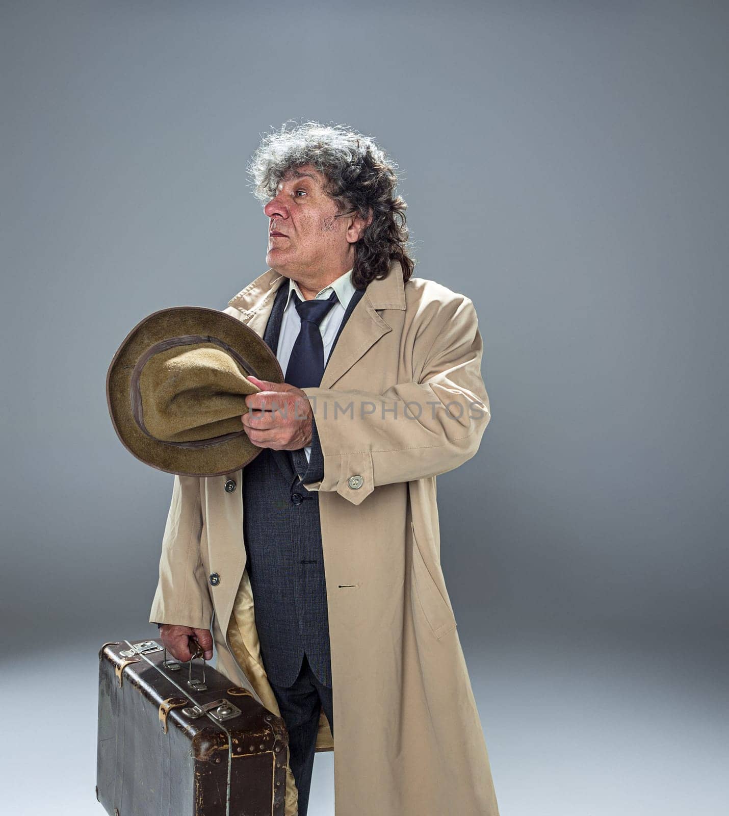 The senior man in cloak as detective or mafia boss. Studio shot on gray in retro stile. Mature man with hat and suitcase