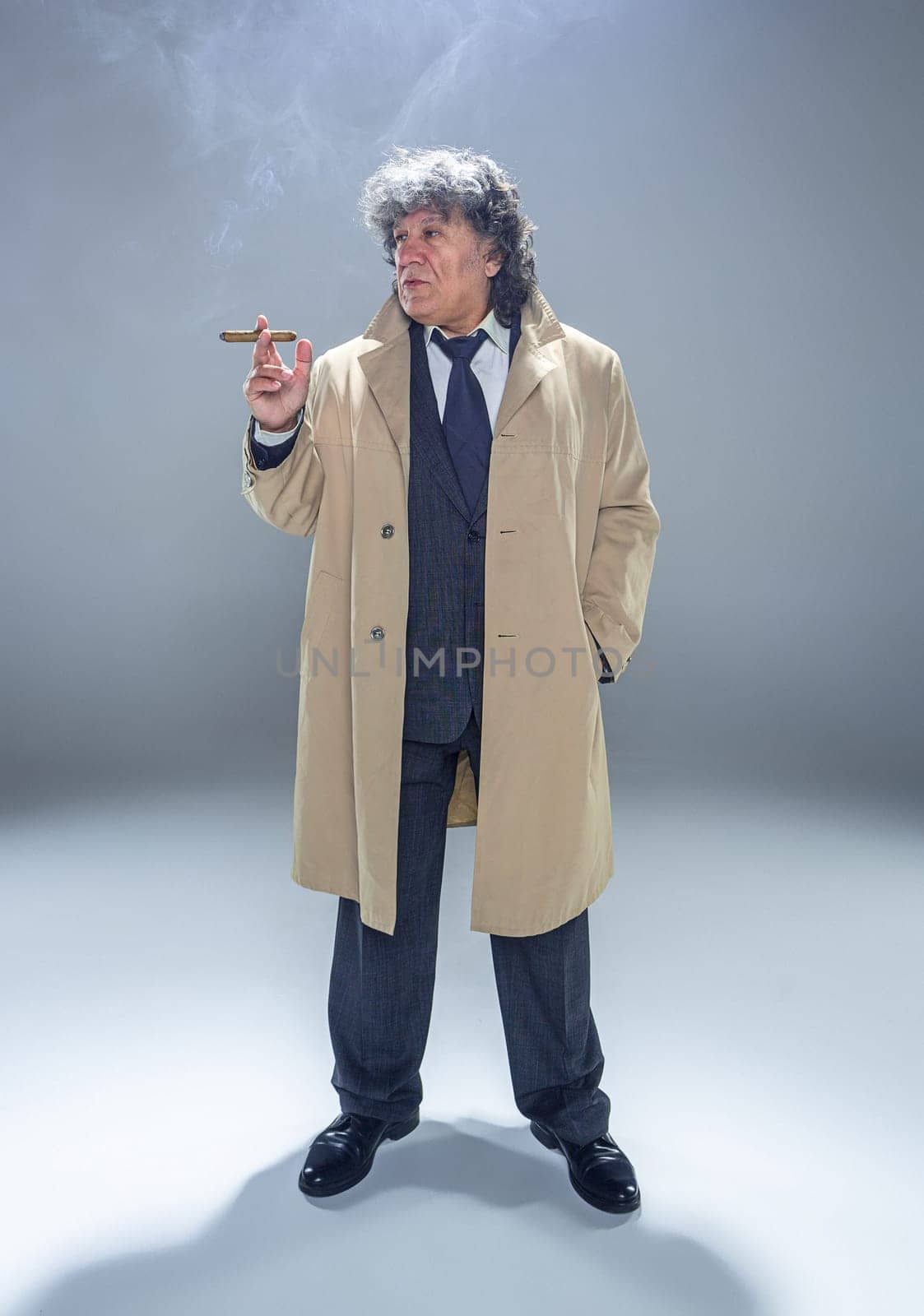 The senior man in cloak with cigar as detective or mafia boss. Studio shot on gray in retro stile