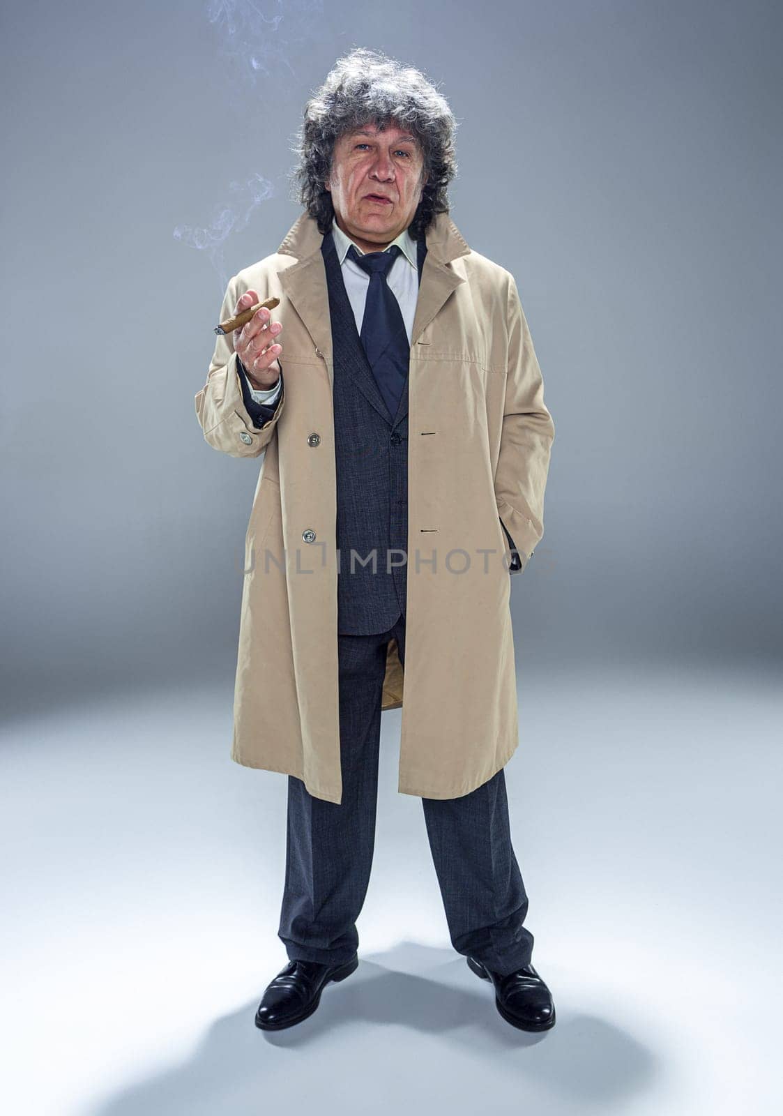 The senior man in cloak with cigar as detective or mafia boss. Studio shot on gray in retro stile