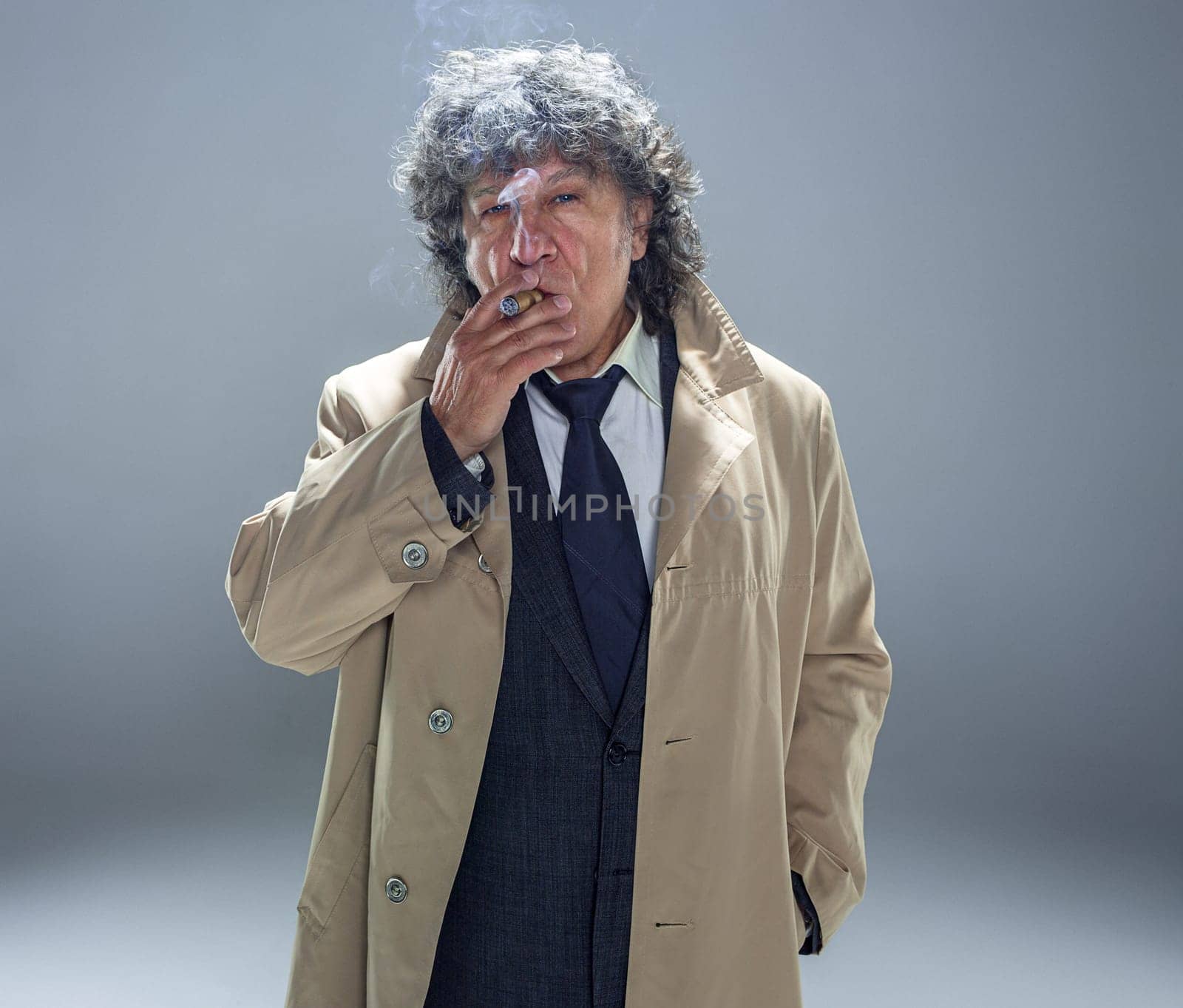 The senior man in cloak with cigar as detective or mafia boss. Studio shot on gray in retro stile