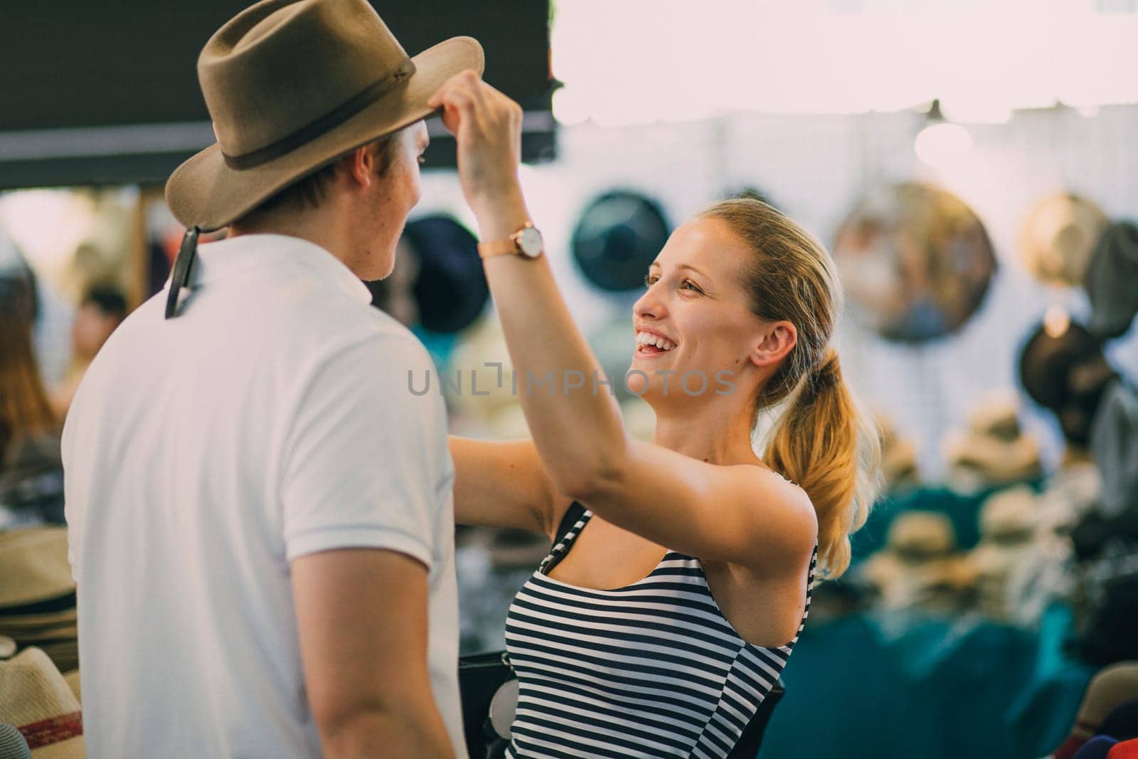 Young couple are exploring Queen Victoria Market in Australia. The woman is putting a hat on her boyfriend in a market stall.