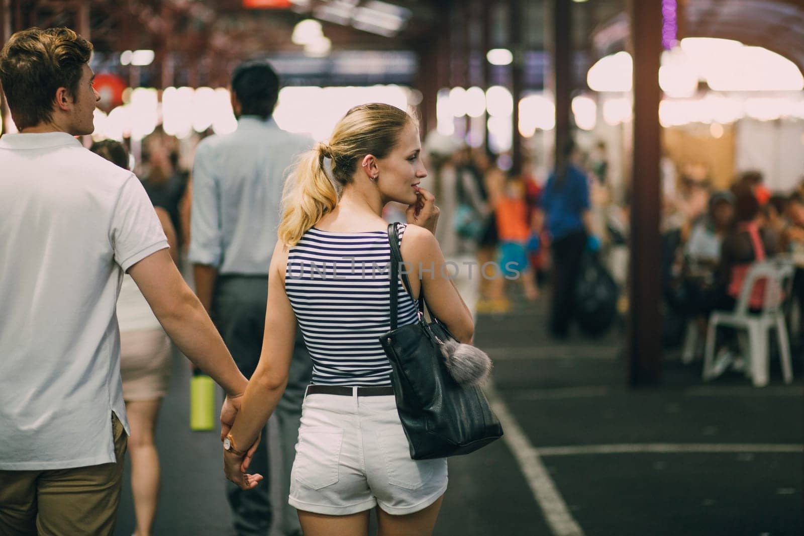 Young couple are exploring Queen Victoria Market in Australia. They are holding hands and the woman is looking out oft he frame to something she is tempted to buy.