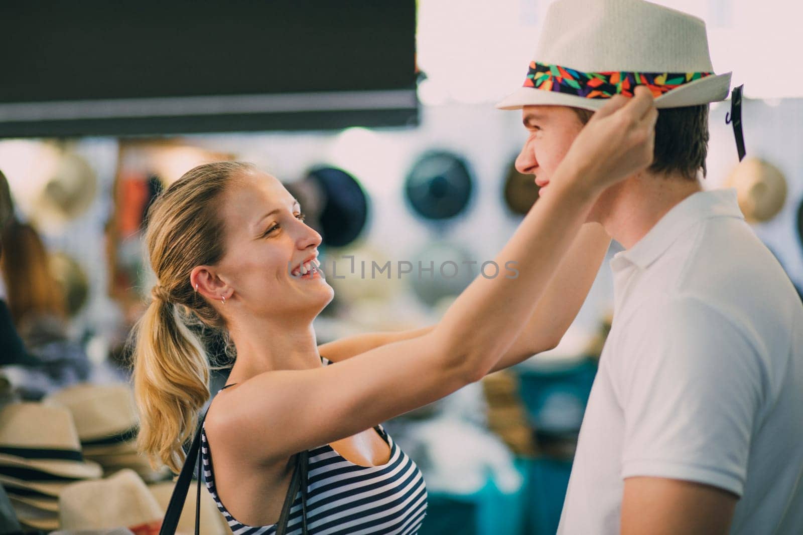 Happy young couple are exploring Queen Victoria Market in Australia. The woman has found a childs summer hat and is trying to squeeze it on her boyfriends head, as he pulls a face.