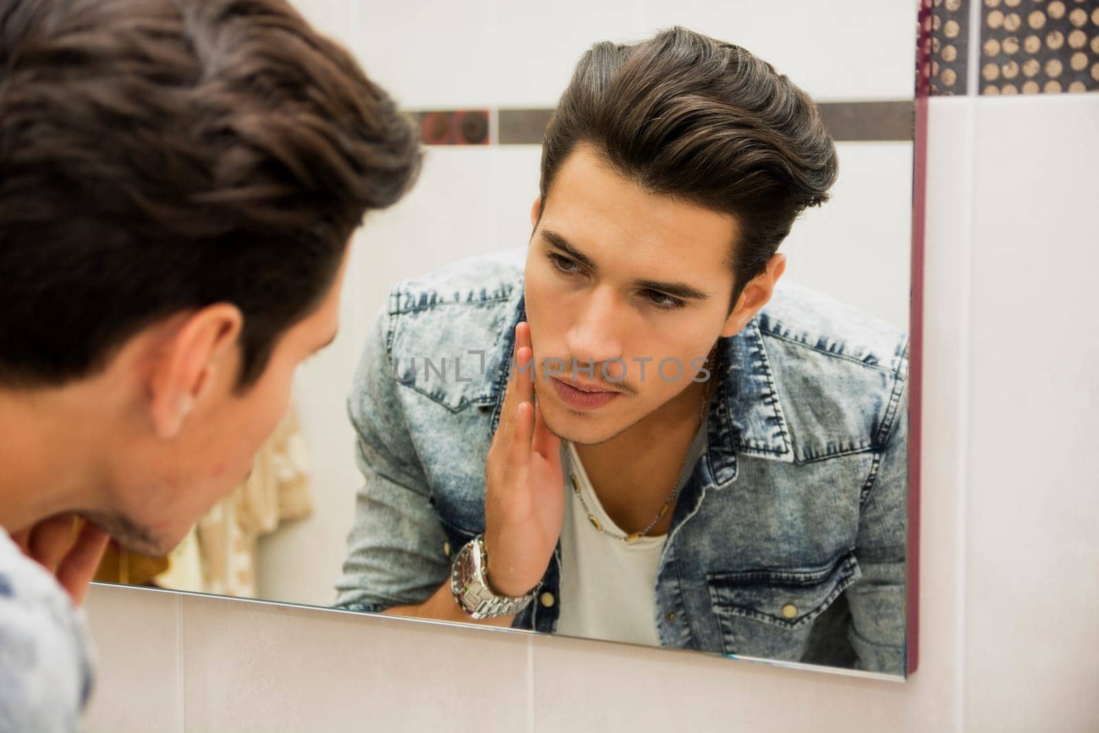Handsome young man examining his face skin up-close, in bathroom mirror in the morning