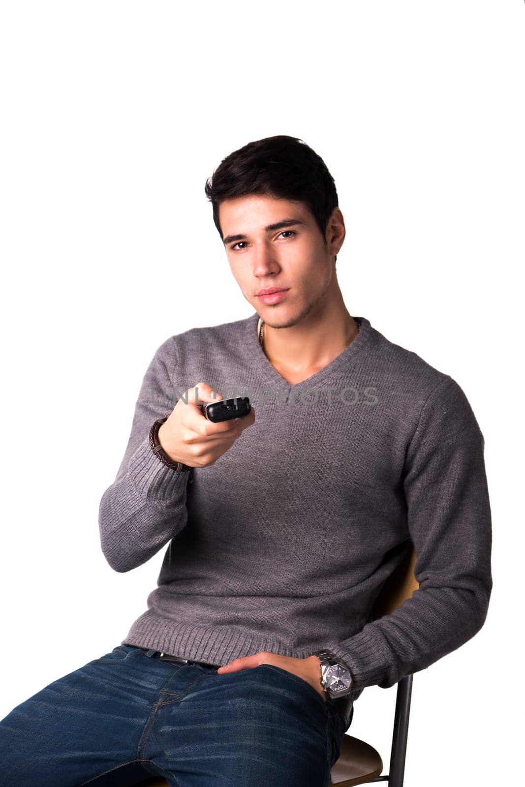 Handsome young man holding remote control and changing TV channels, sitting isolated on white